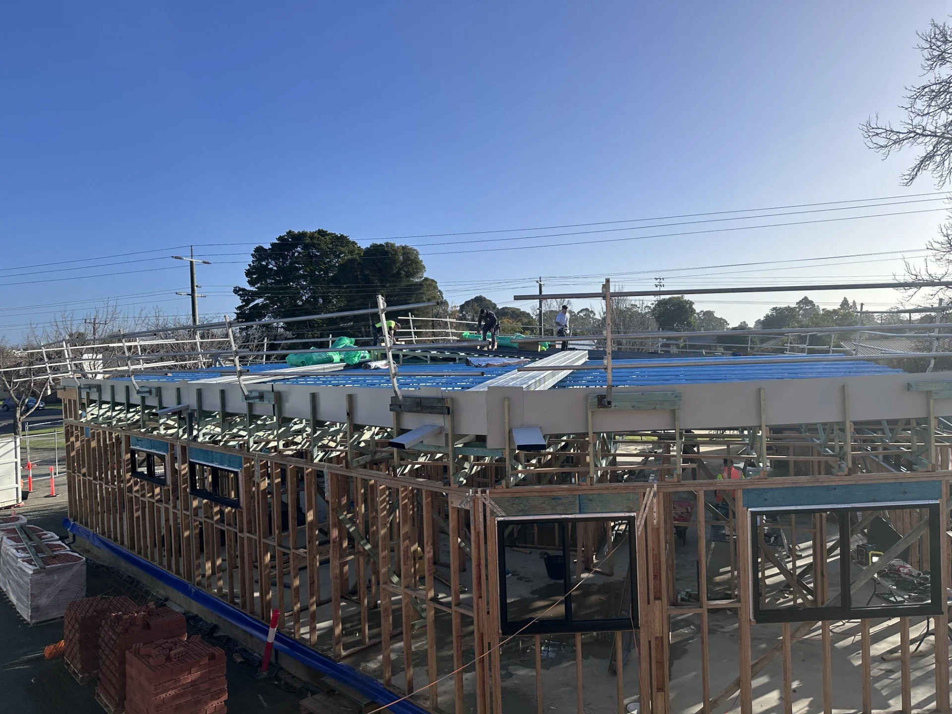 Construction site of a building with a blue roof, workers are on the roof installing materials. The building's wooden framework is visible, and there are piles of bricks and construction materials on the ground. Power lines and trees are in the background under a clear blue sky.