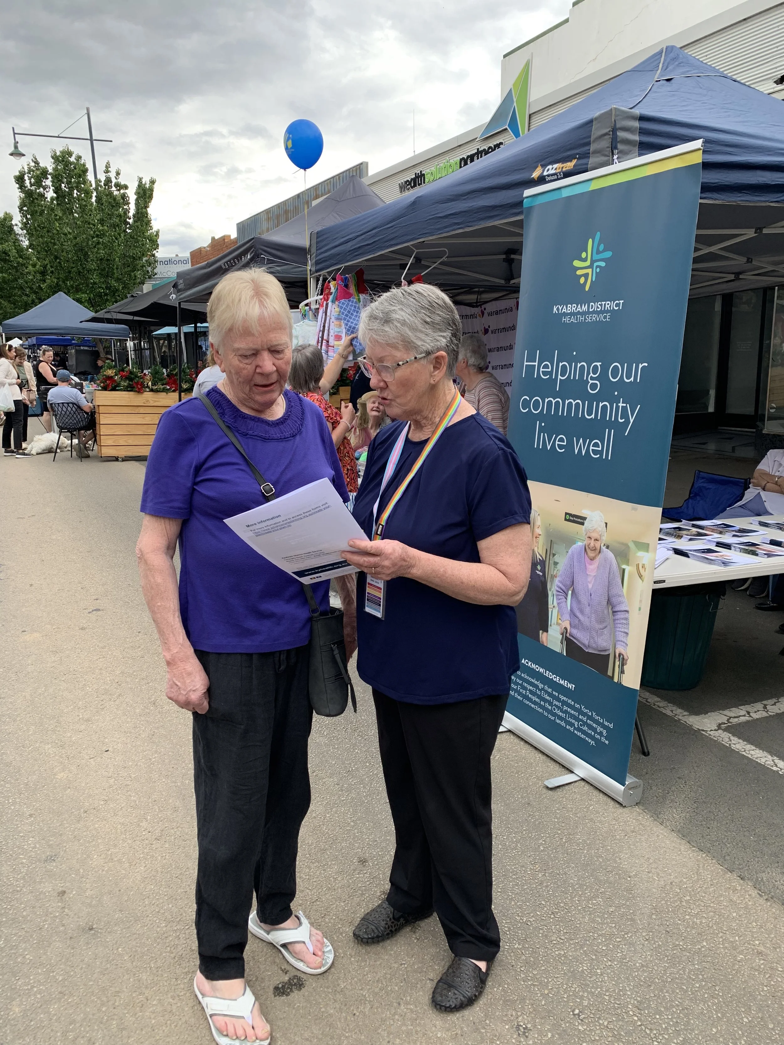 Two elderly women standing outdoors at a community event, reading a piece of paper together near a Kyabram District Health Service banner, with tents and other people visible in the background.