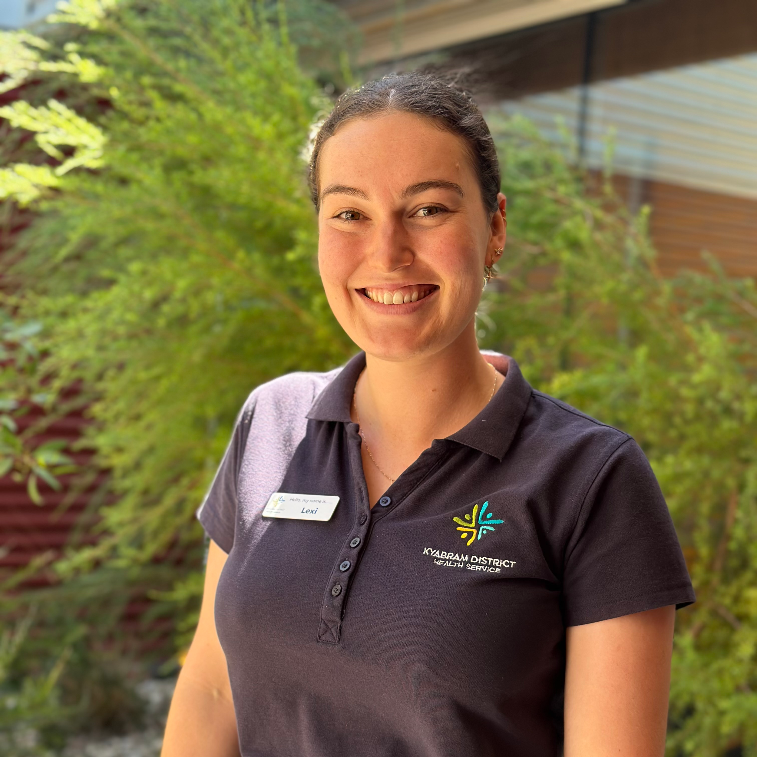 Close-up of a smiling woman wearing a black polo shirt with a name tag, standing outdoors with greenery and a wooden structure in the background.