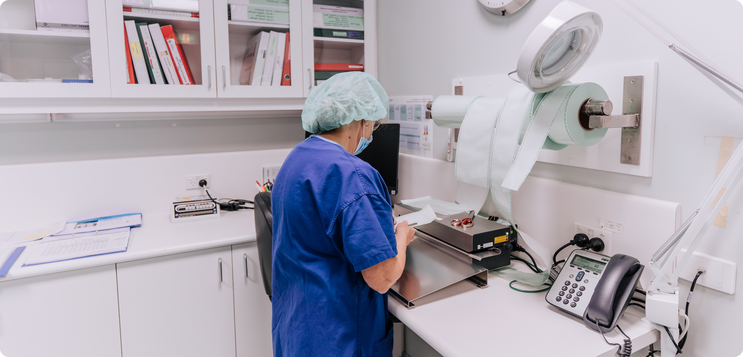 A scientist wearing scrubs, a hair cover, and a face mask is working with a roll of medical tape or sterile strips in a laboratory or medical setting. There are cabinets with binders and lab equipment in the background.