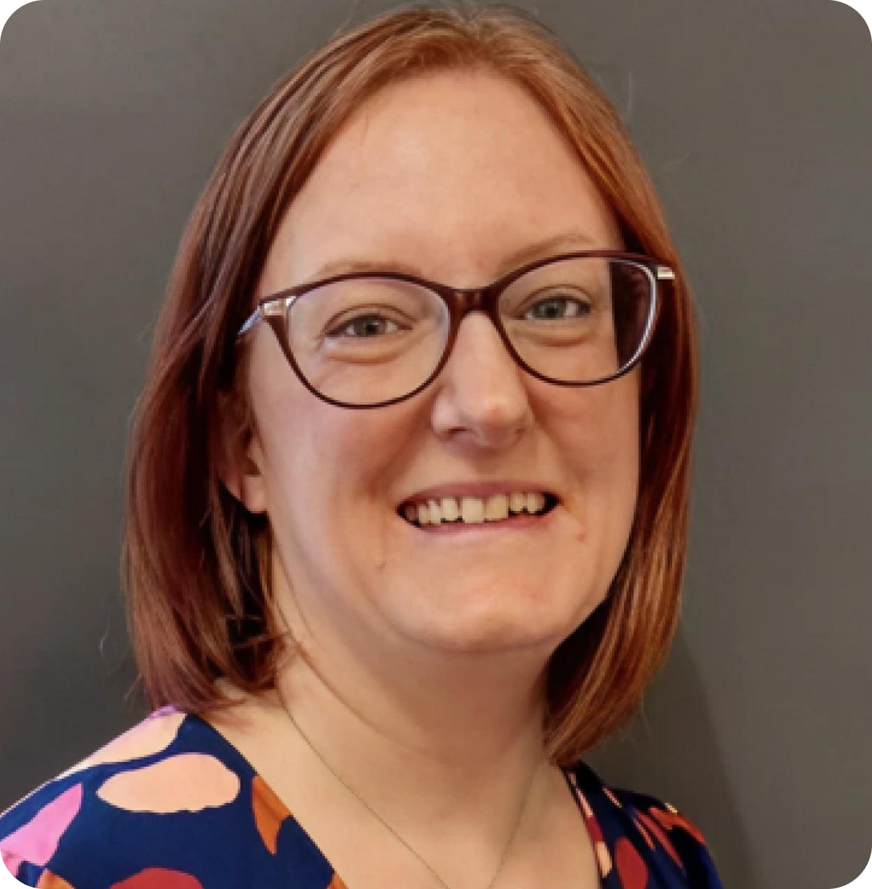 Close-up of a woman with shoulder-length red hair, wearing glasses, smiling, against a plain gray background.