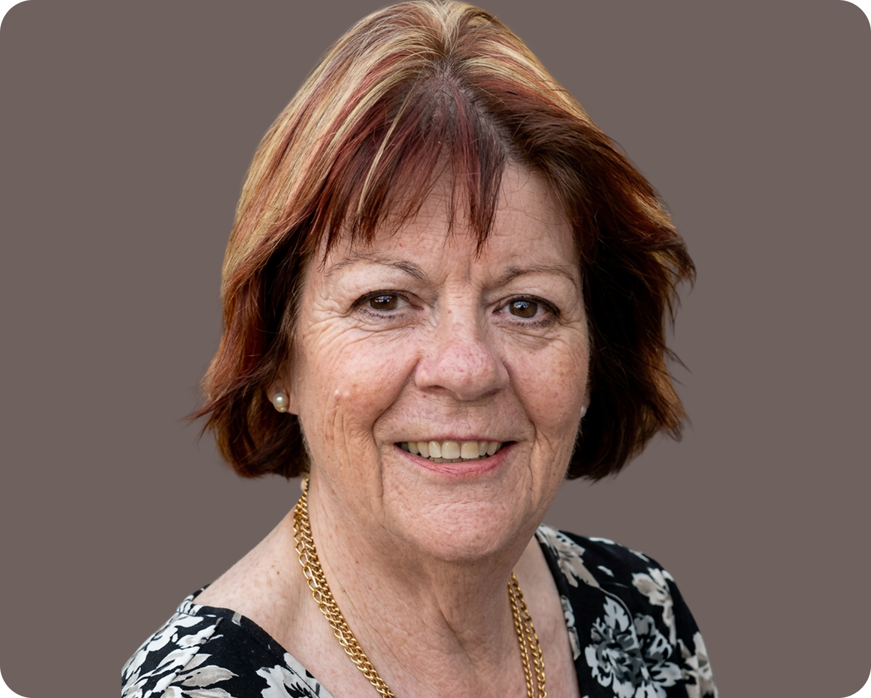A smiling middle-aged woman with short red hair, wearing pearl earrings, a black and white floral top, gold necklace, and a grey background.