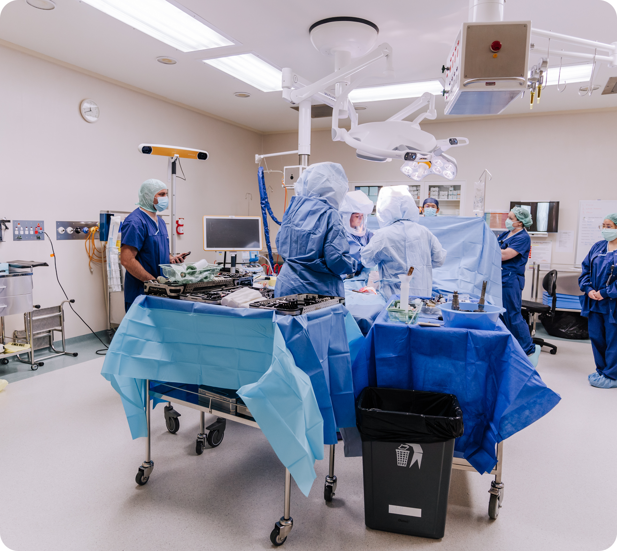 Hospital operating room with medical staff dressed in scrubs and masks, surgical equipment, monitors, and overhead surgical lights.