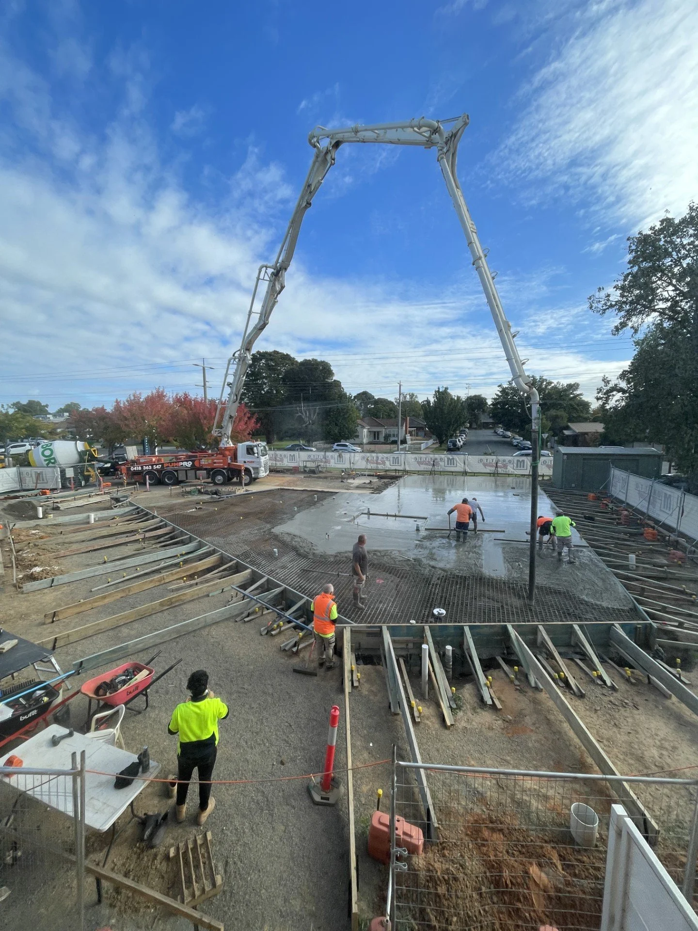 Construction workers pouring concrete on a building foundation with a concrete pump truck overhead and partially framed concrete slabs around the site.