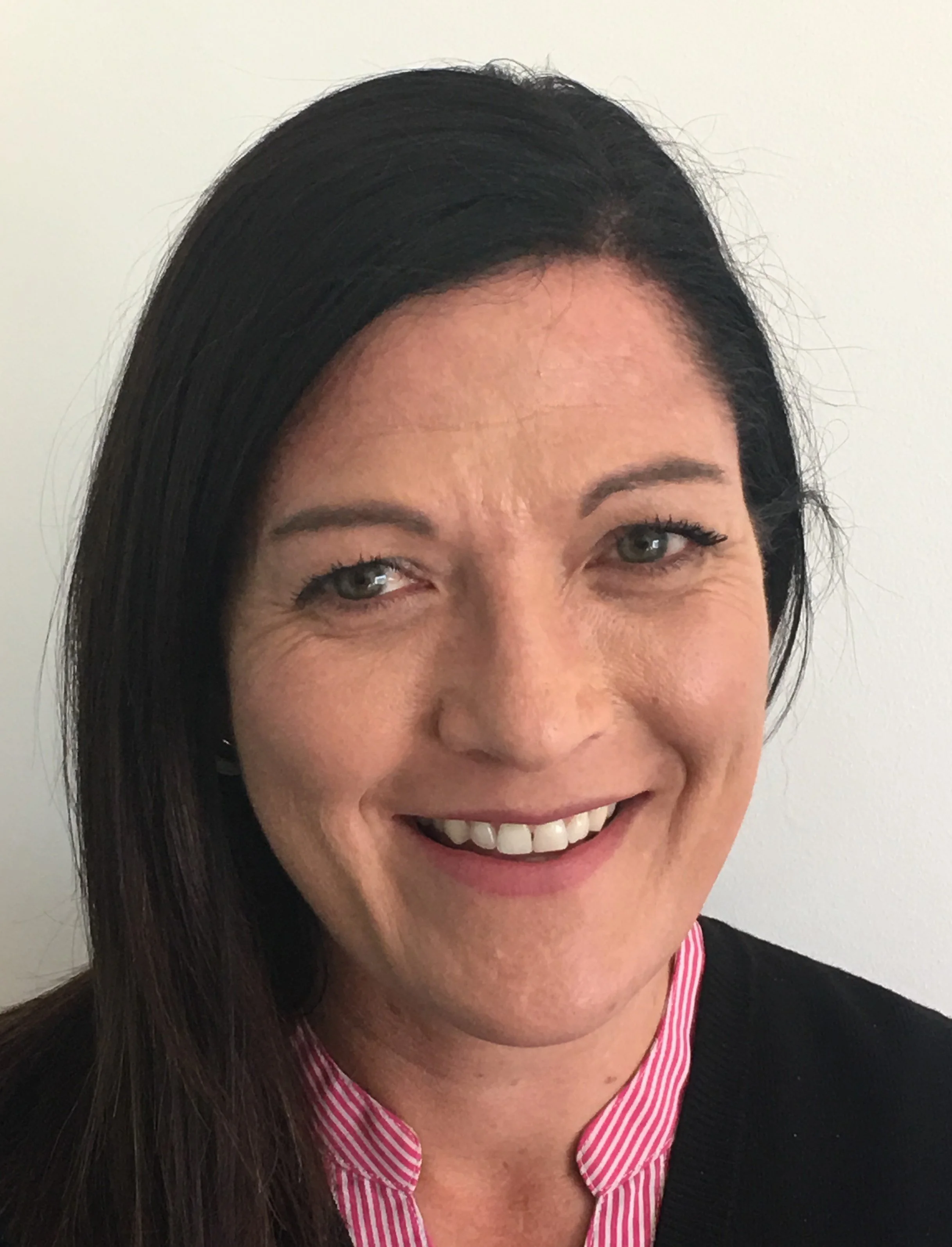 Close-up of a smiling woman with dark hair, wearing a pink and white striped shirt under a black sweater, against a plain white background.