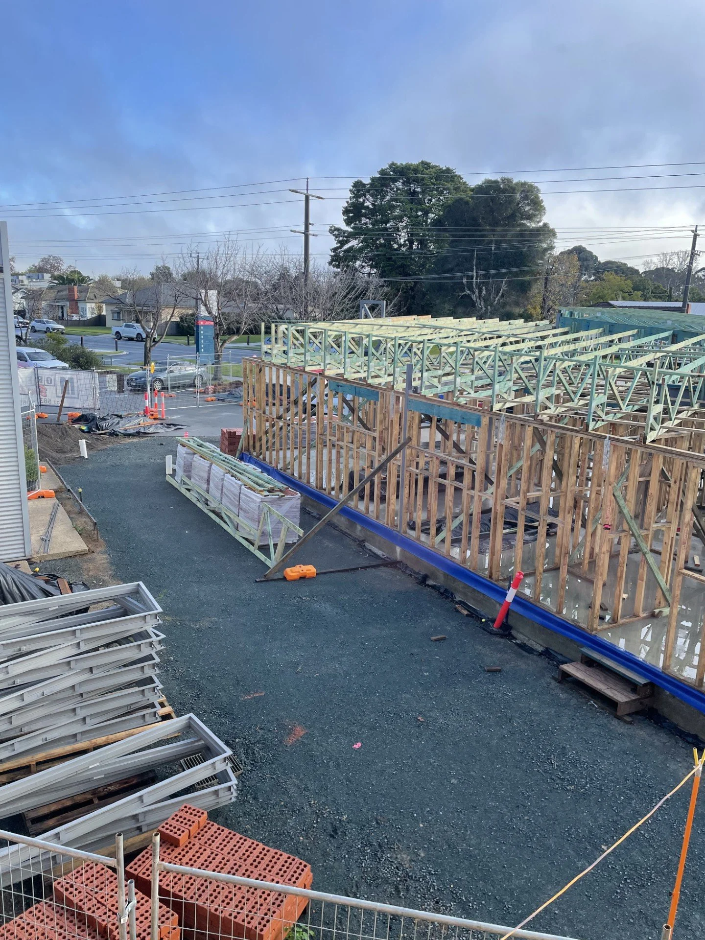 Construction site with wooden framing for building, orange safety cones, and building materials on the ground, outdoor setting with trees and parked cars in the background.