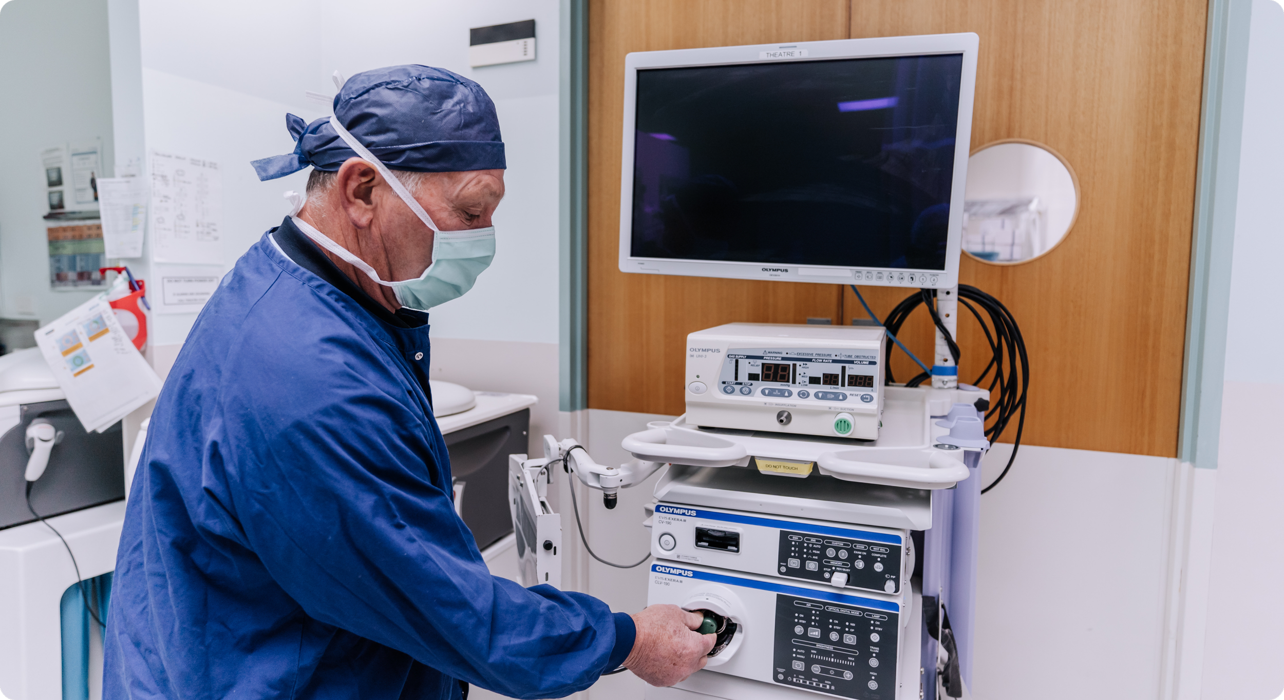A healthcare professional in scrubs, surgical cap, and mask working with medical equipment in a hospital room.