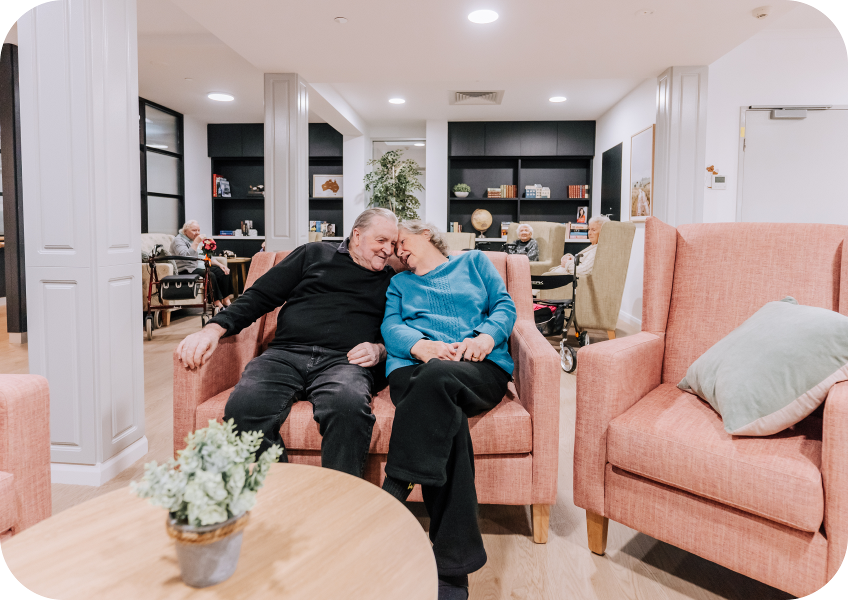 An elderly man and woman are sitting together on a pink sofa in a care home common area, leaning heads together and smiling, with other elderly residents in the background on chairs and wheelchairs.