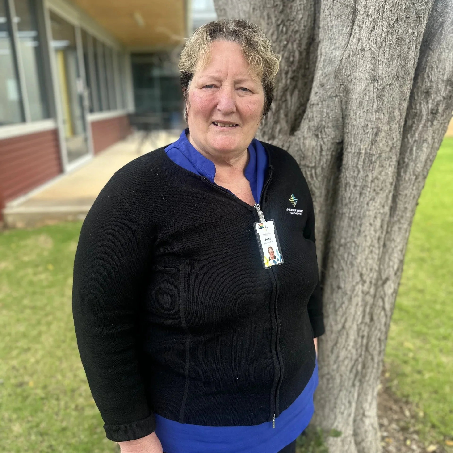 Close-up of a middle-aged woman with short wavy hair standing outdoors next to a large tree trunk, wearing a black jacket with a logo and a blue shirt underneath, and a name badge, smiling at the camera.