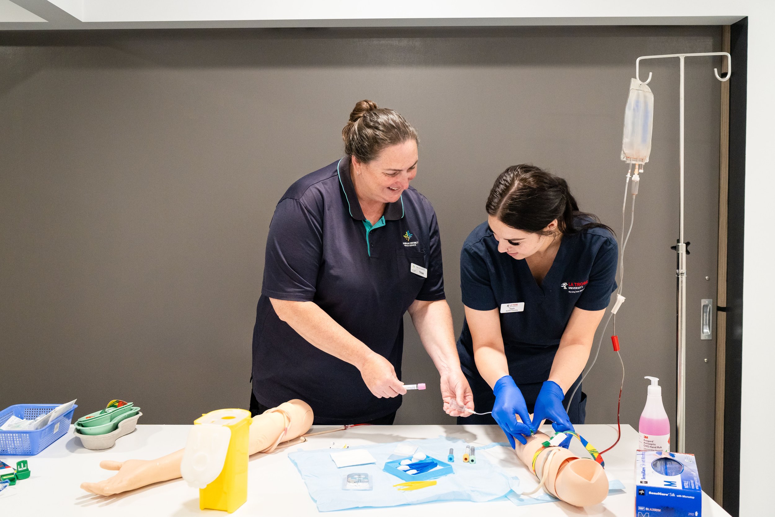 Two women in medical scrubs practicing a medical procedure on a training arm with a simulated IV line, various medical supplies on the table, and an IV stand nearby.
