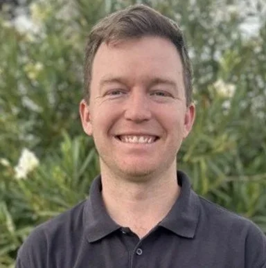 A smiling man with short brown hair and a black collared shirt standing outdoors in front of green foliage.