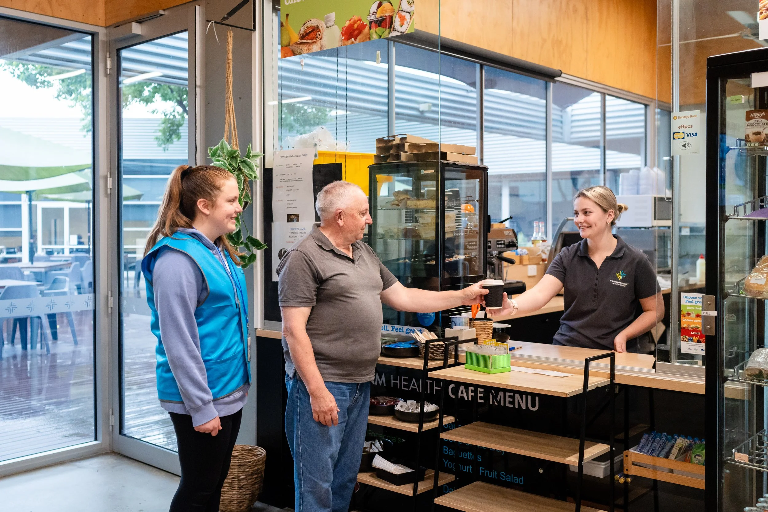 A man is purchasing a drink from a female employee at a cafe counter. A young woman is standing nearby inside the cafe, and large glass windows reveal an outdoor seating area and trees outside.