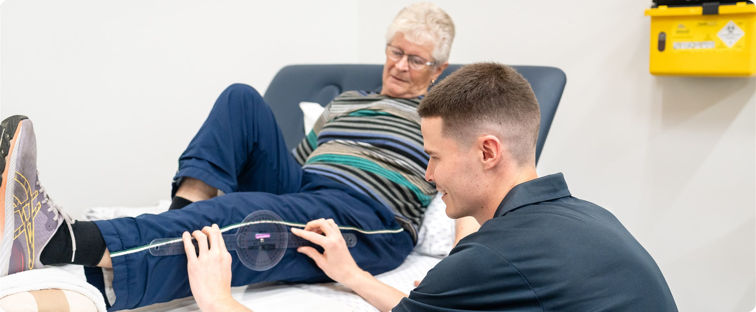 Young man receiving a blood test from a healthcare professional while an older woman lies on a hospital bed.