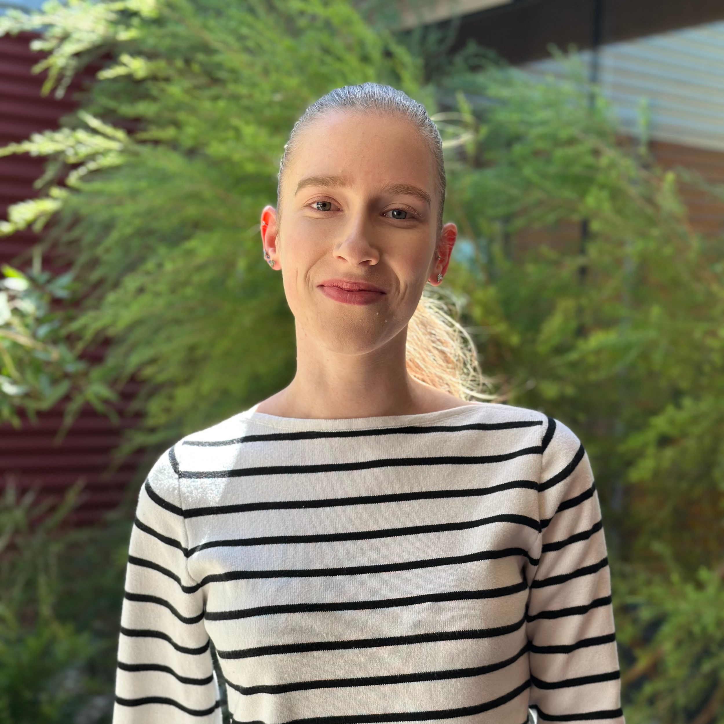 Close-up of a young woman with light skin, blonde hair in a ponytail, wearing earrings, and sporting a white and black striped shirt, smiling outdoors with green foliage and a building in the background.