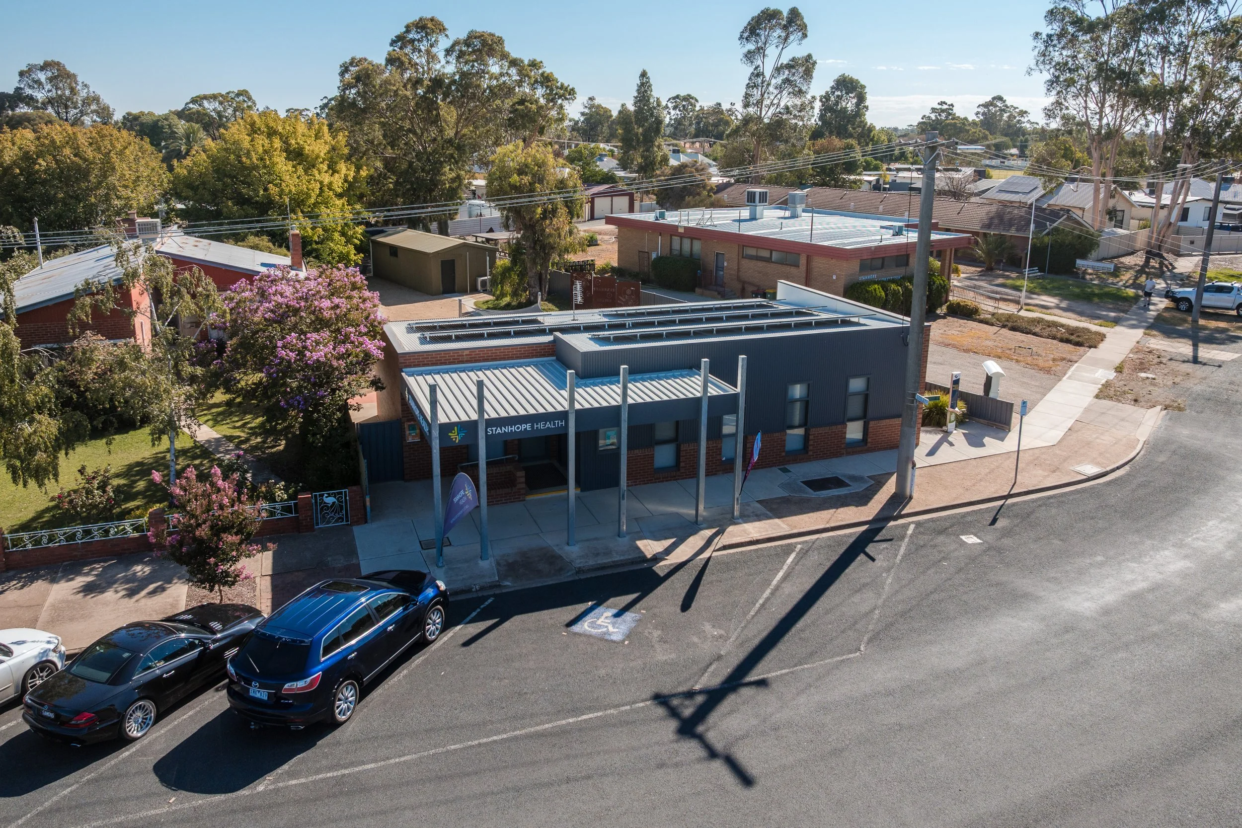 An aerial view of a small urban area with cars parked along the street, including a designated handicapped parking space, a building with a sign for Stanhope Health, surrounded by trees and residential houses.