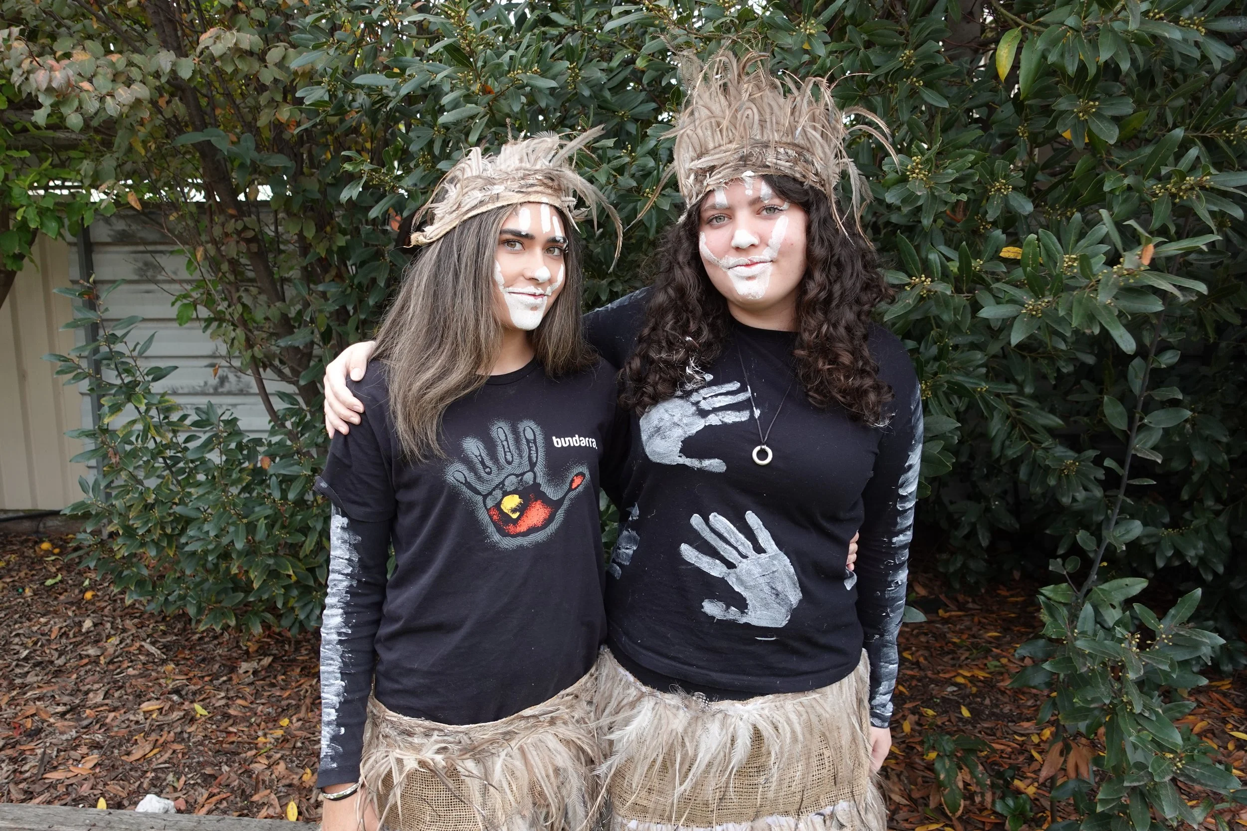 Two young women standing outdoors in front of dense green foliage, dressed in tribal or cultural costumes with face paint, feathered headpieces, and skirts made from natural materials.