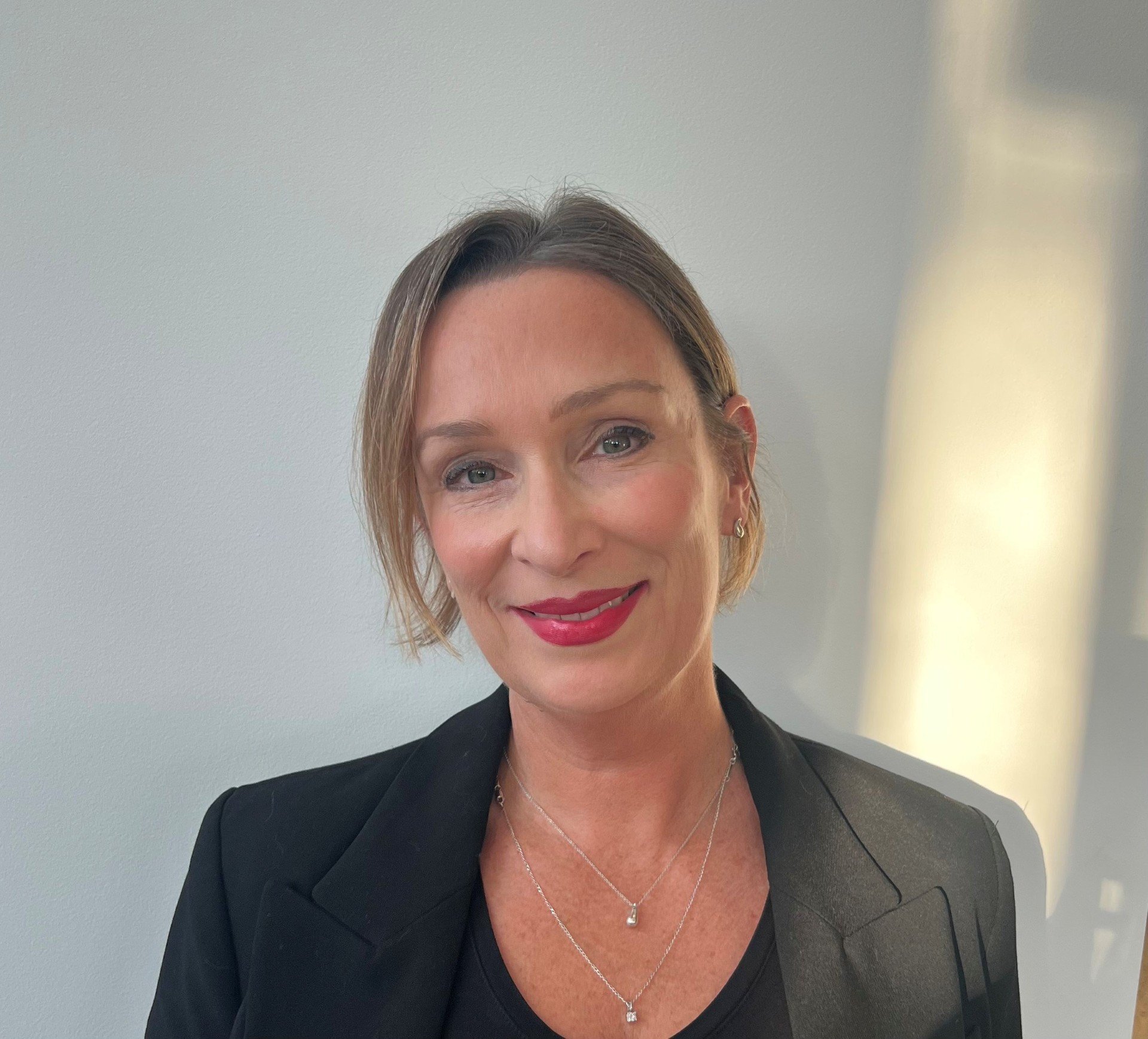 Close-up of a woman with short light brown hair, wearing makeup, a black blazer, and silver jewelry, smiling slightly, with a plain light-colored background.