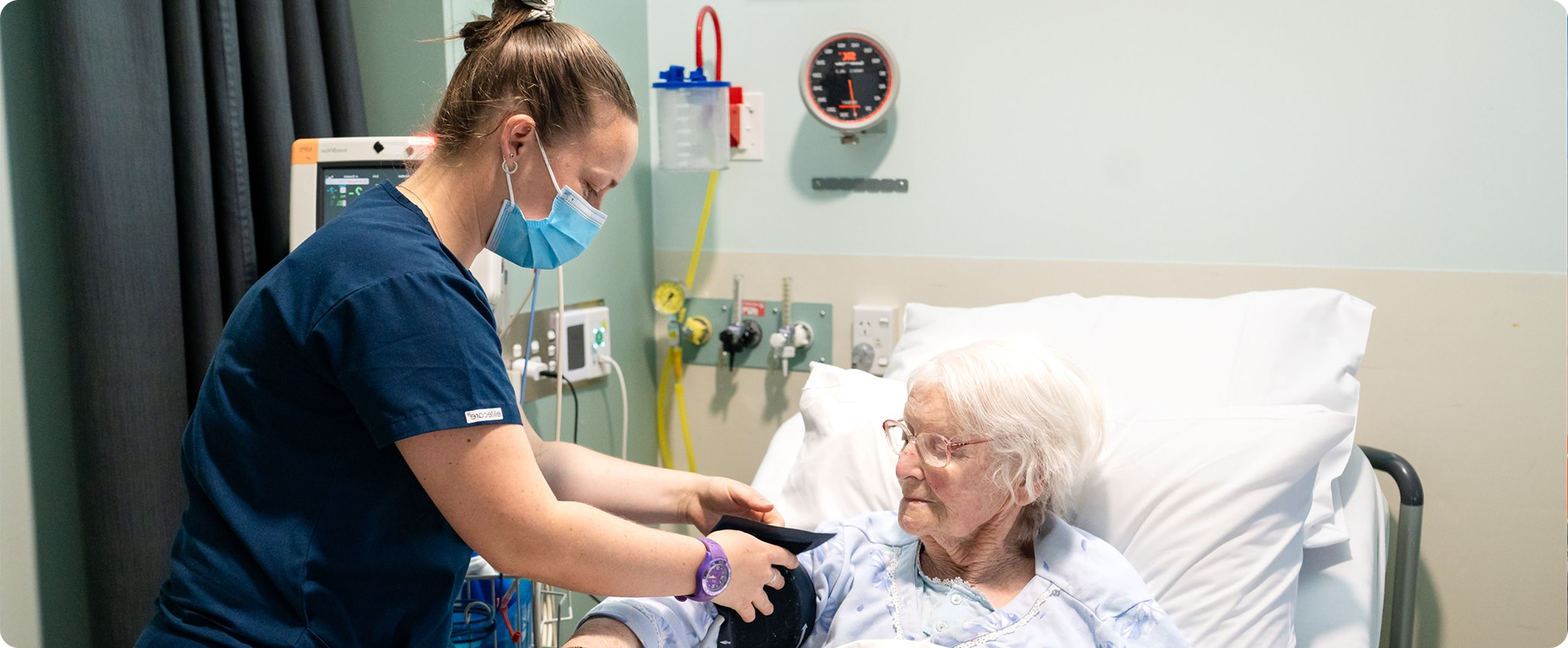 A nurse in blue scrubs, wearing a face mask, checks the blood pressure of an elderly woman lying in a hospital bed. The woman has gray hair, glasses, and a hospital gown, and is looking at the nurse as she takes her blood pressure.