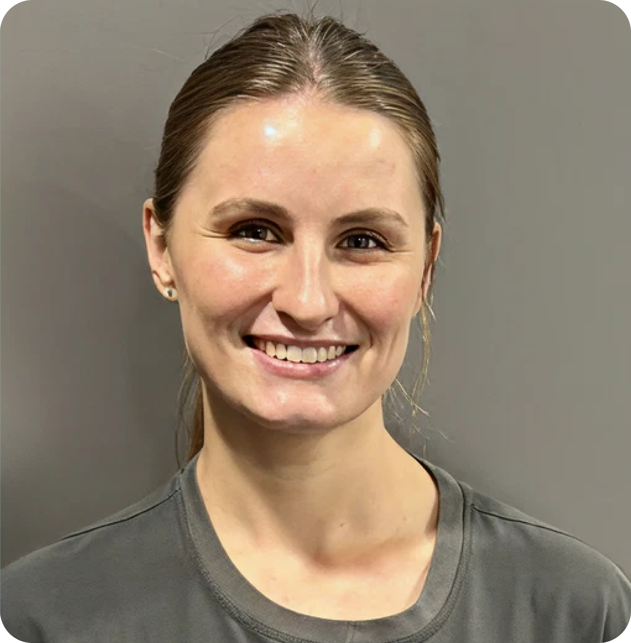 A smiling woman with light brown hair tied back, wearing a dark gray t-shirt, standing against a plain gray background.