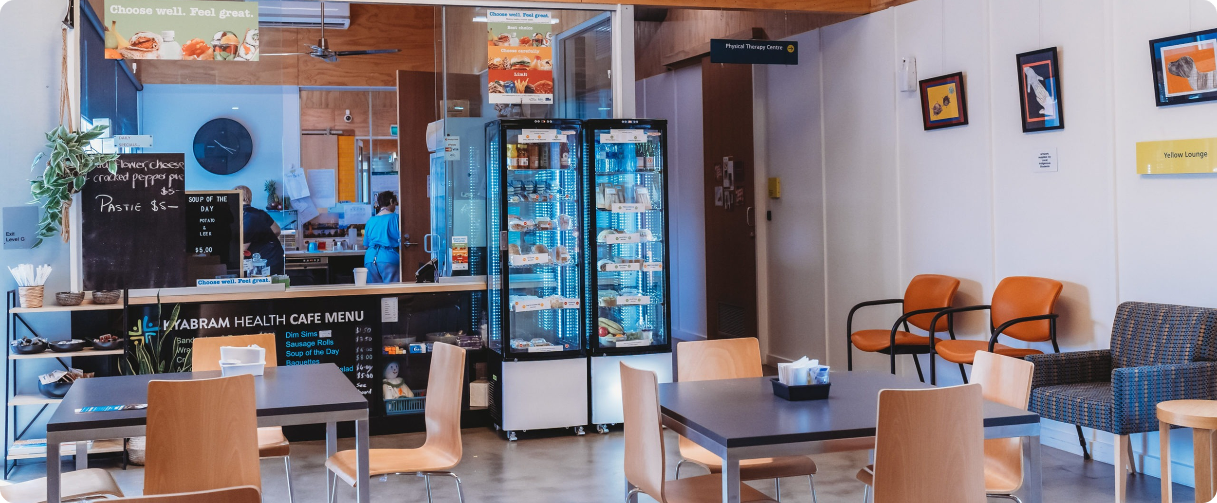 Interior of a cafe with tables, chairs, a glass display refrigerator, and a counter area with a staff member. There are framed pictures and signs on the walls, and a sign indicating a physical therapy center. The cafe menu is visible.
