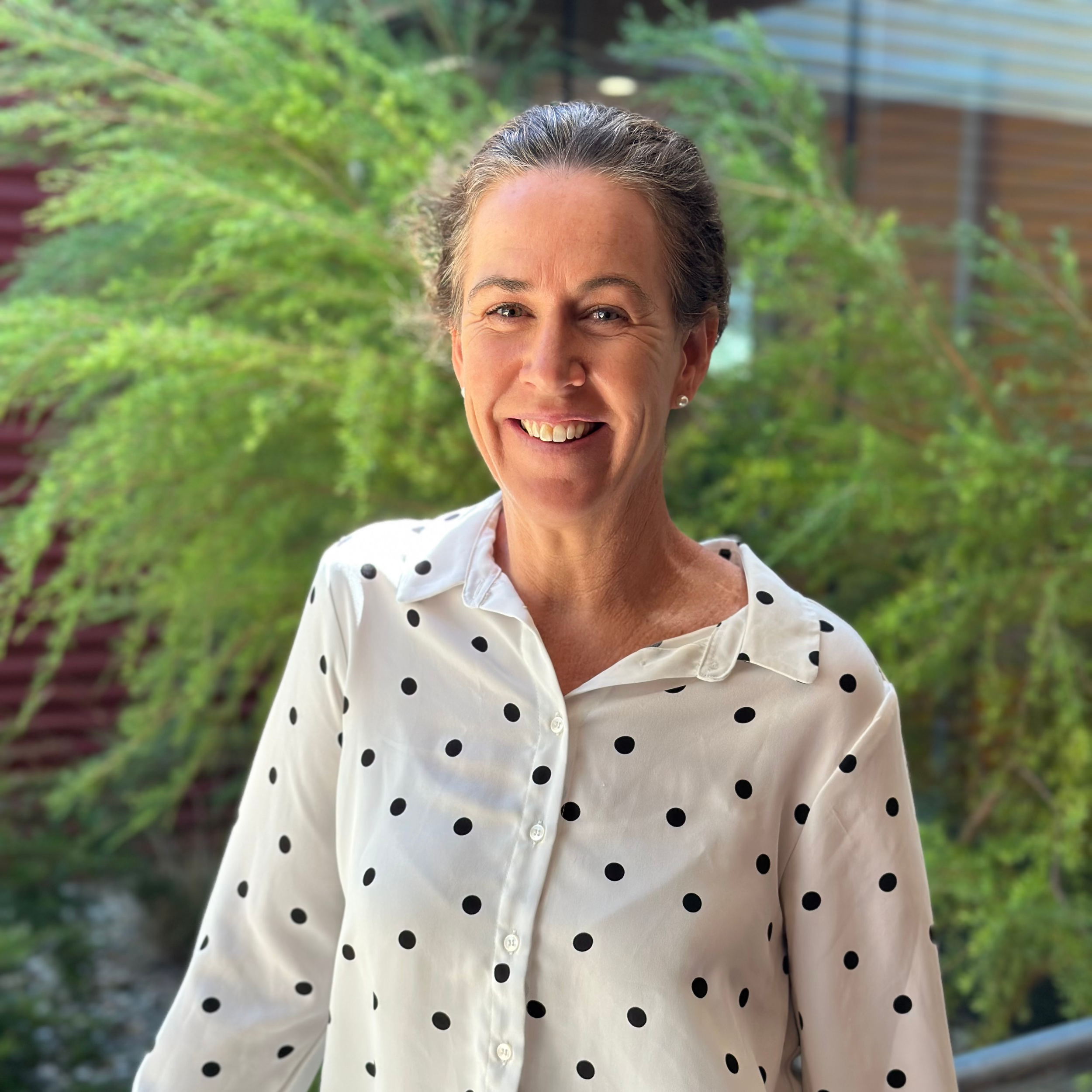 A smiling woman with gray hair styled back, wearing a white blouse with black polka dots, standing outdoors with green foliage and a building with horizontal siding in the background.