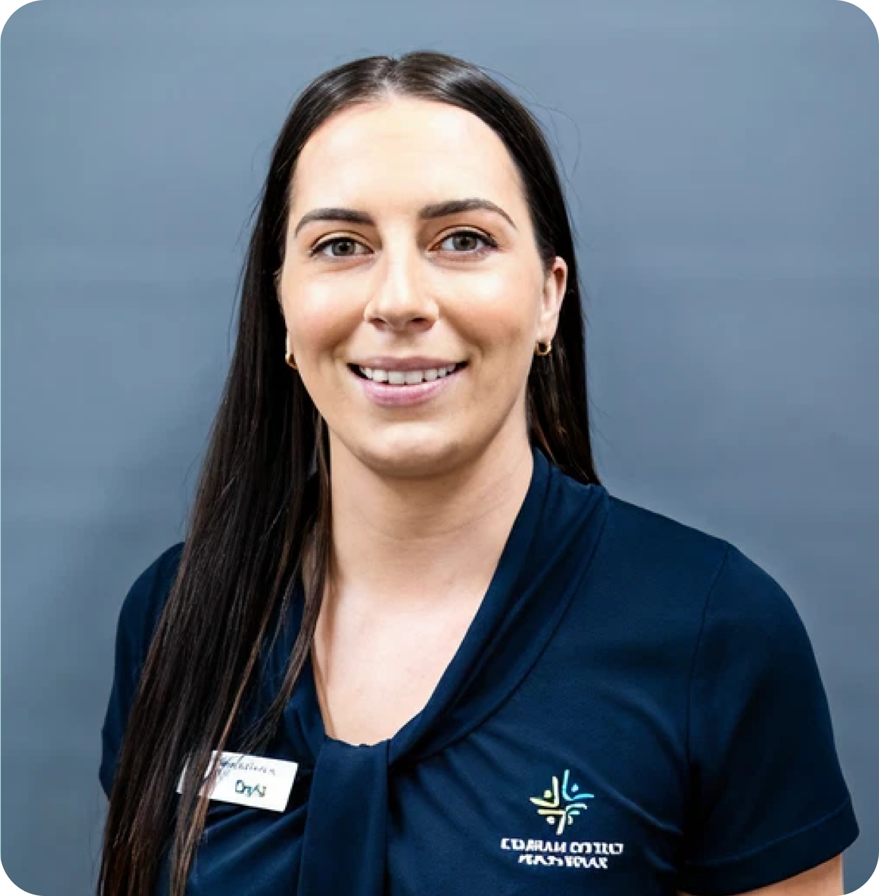 A smiling woman with long dark hair wearing a navy blue shirt with a logo and a name tag, standing against a gray background.