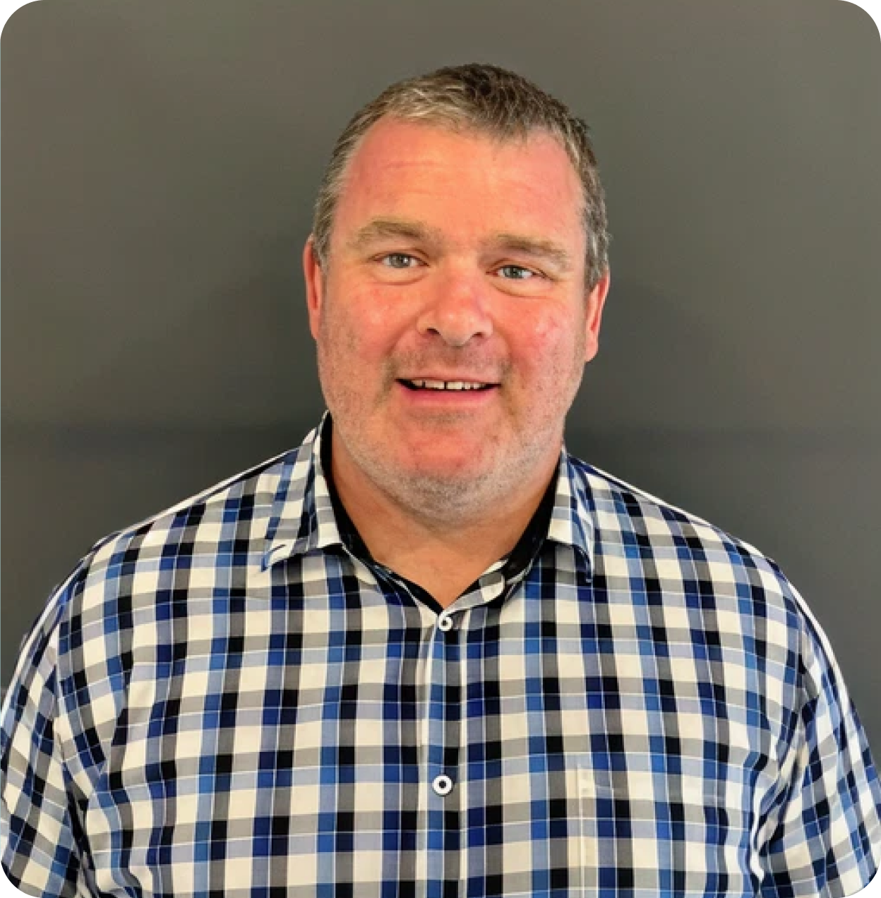 Portrait of a man with short gray hair, wearing a blue and white checkered shirt, smiling in front of a plain gray background.