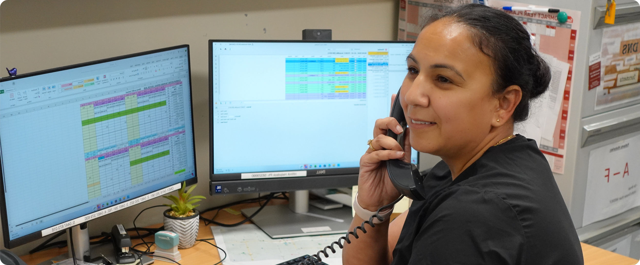 Woman working at a desk with dual computer monitors displaying spreadsheets, talking on a corded phone, with a small potted plant on the desk and various office supplies around.