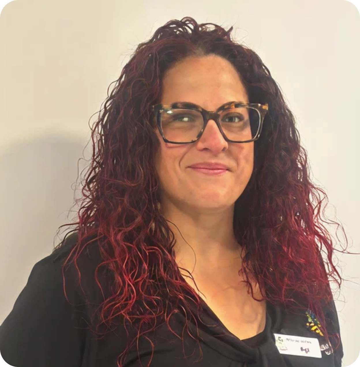 A woman with curly, reddish hair wearing large glasses and a black shirt with a name tag, standing against a plain white background.