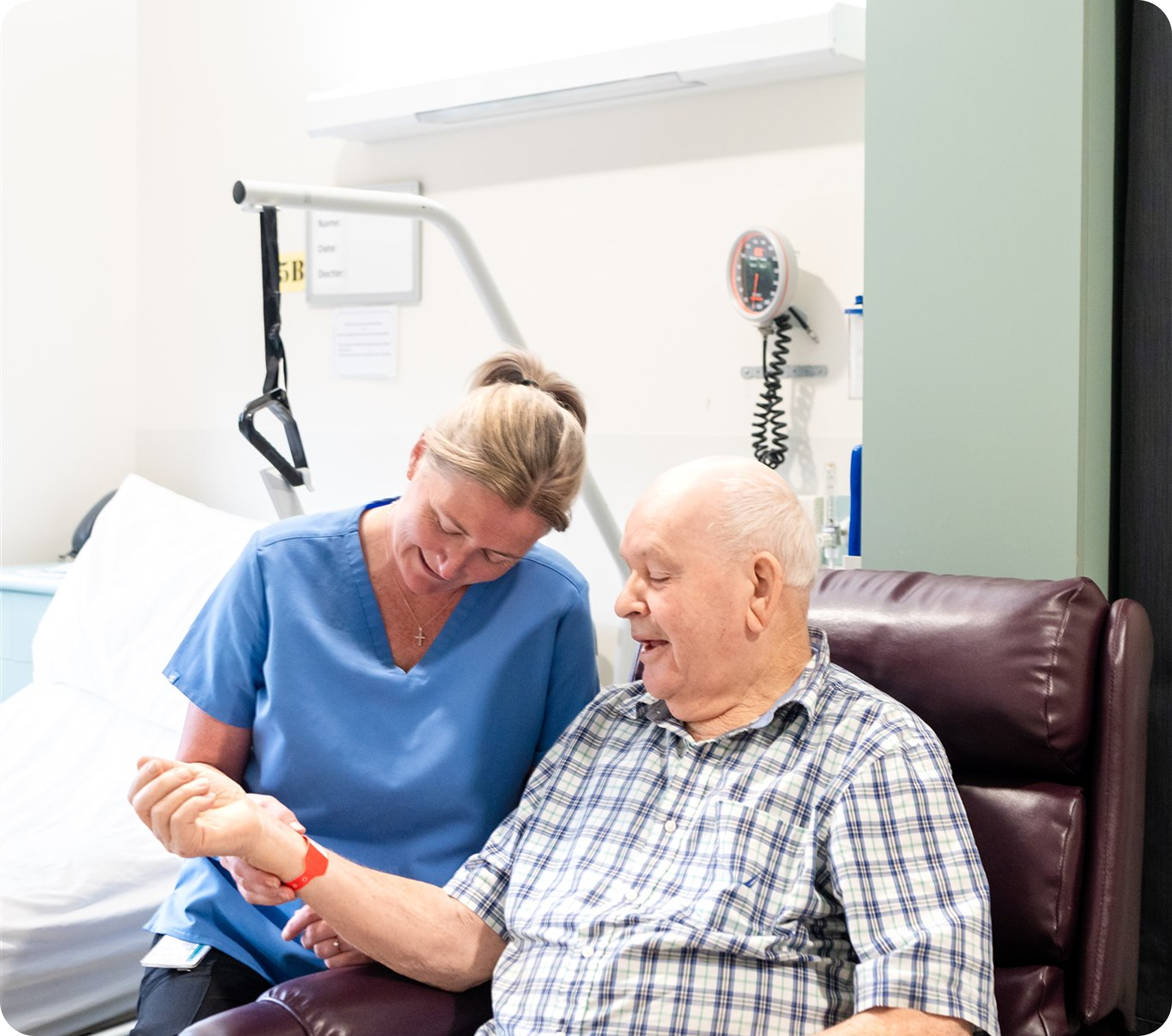 A healthcare worker, wearing blue scrubs, is smiling and kneeling in front of an elderly man in a checkered shirt, both smiling and engaging in a conversation in a hospital room.