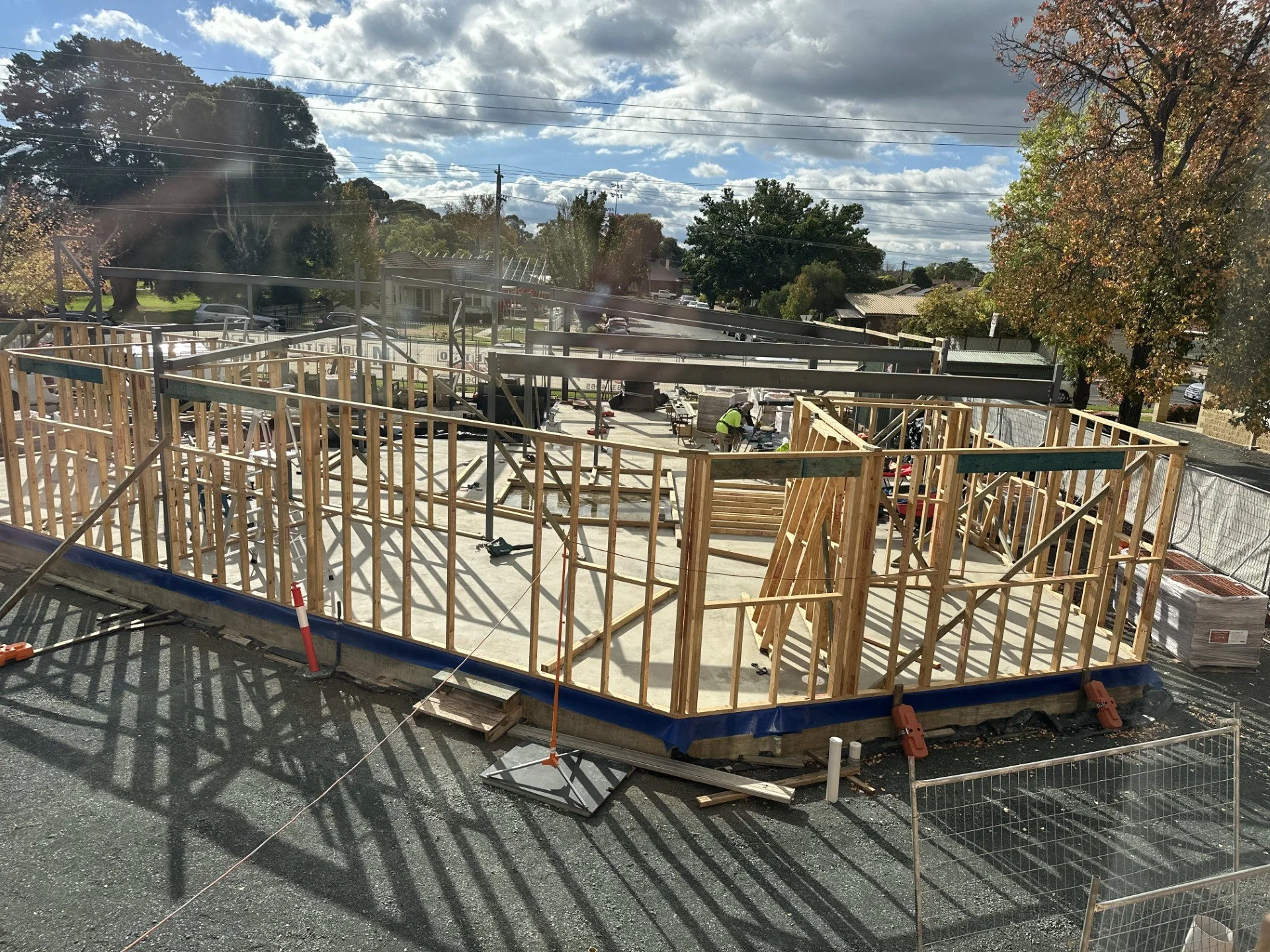 Construction site with wooden framing for a building, workers, and equipment under a partly cloudy sky, surrounded by trees and residential area.