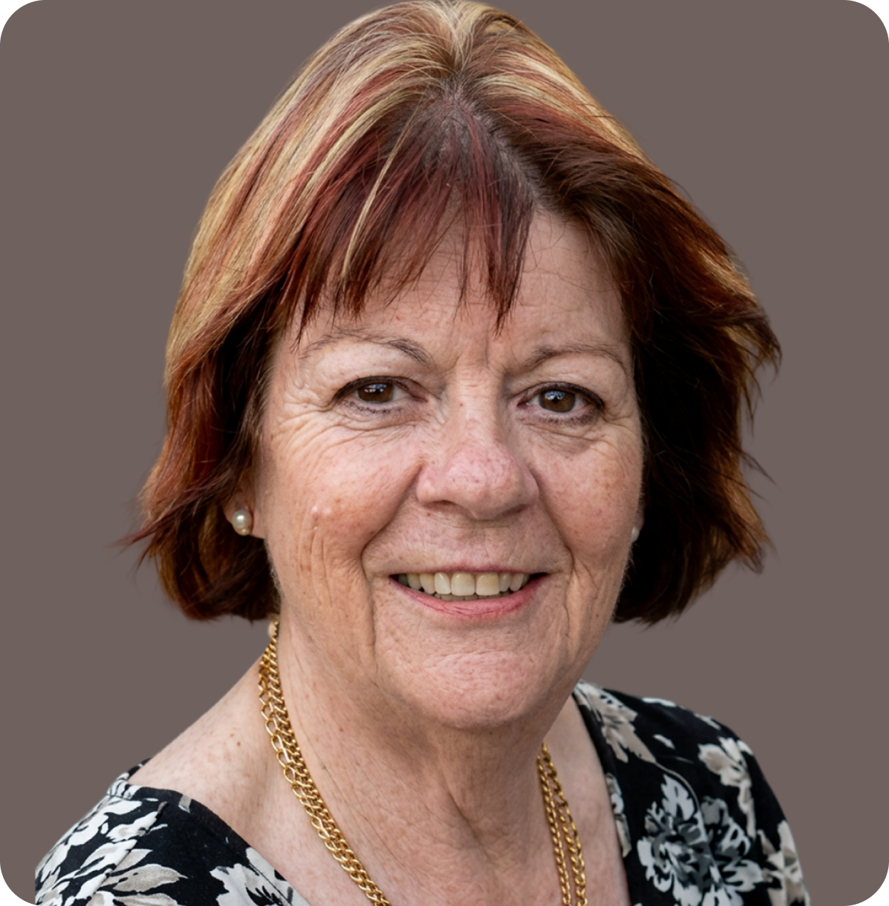 A smiling middle-aged woman with short reddish-brown hair, wearing pearl earrings, a gold chain necklace, and a black and white floral top, against a plain gray background.