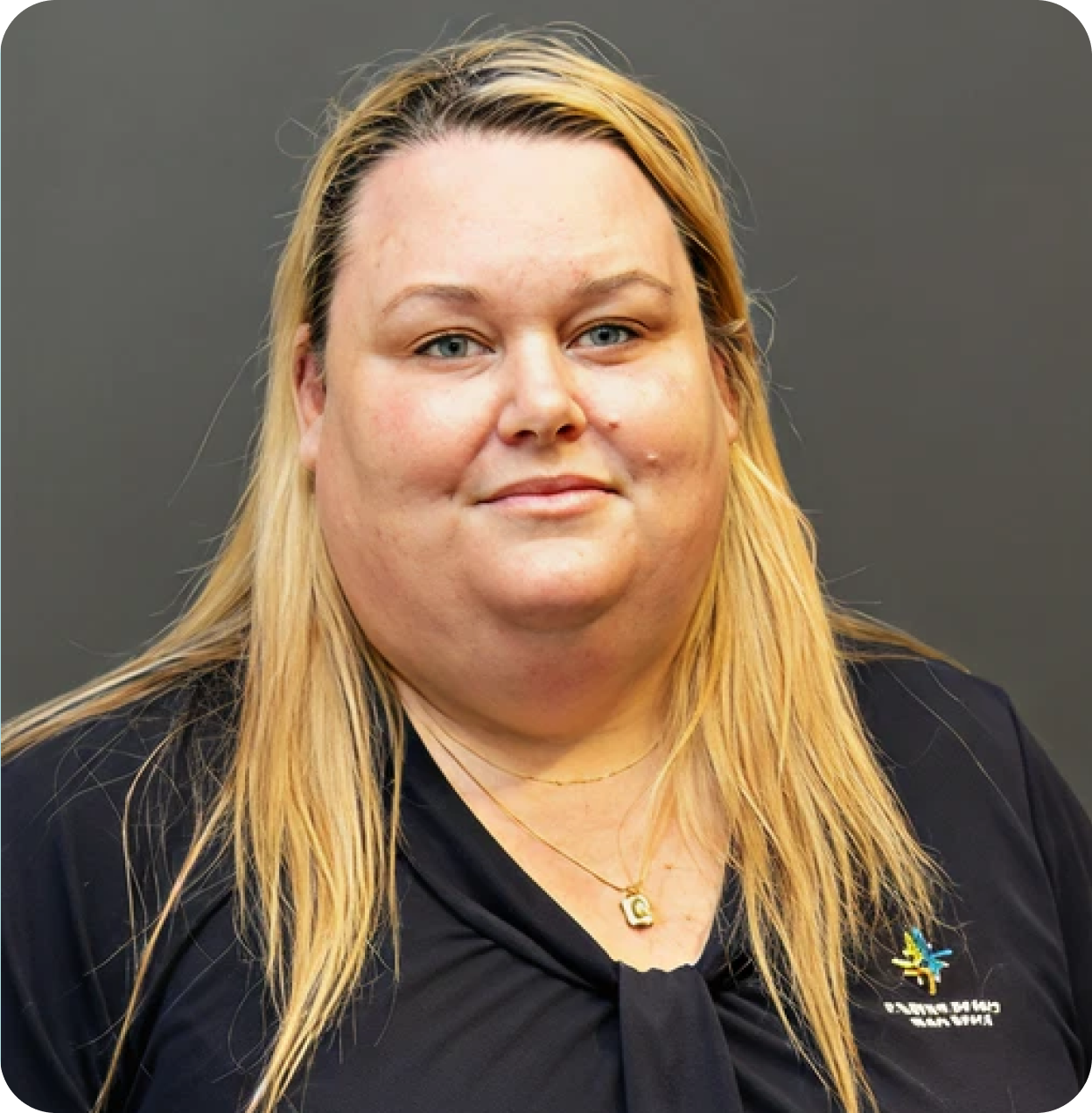 A woman with long blonde hair and blue eyes wearing a black shirt with a logo and a gold necklace with the letter G.