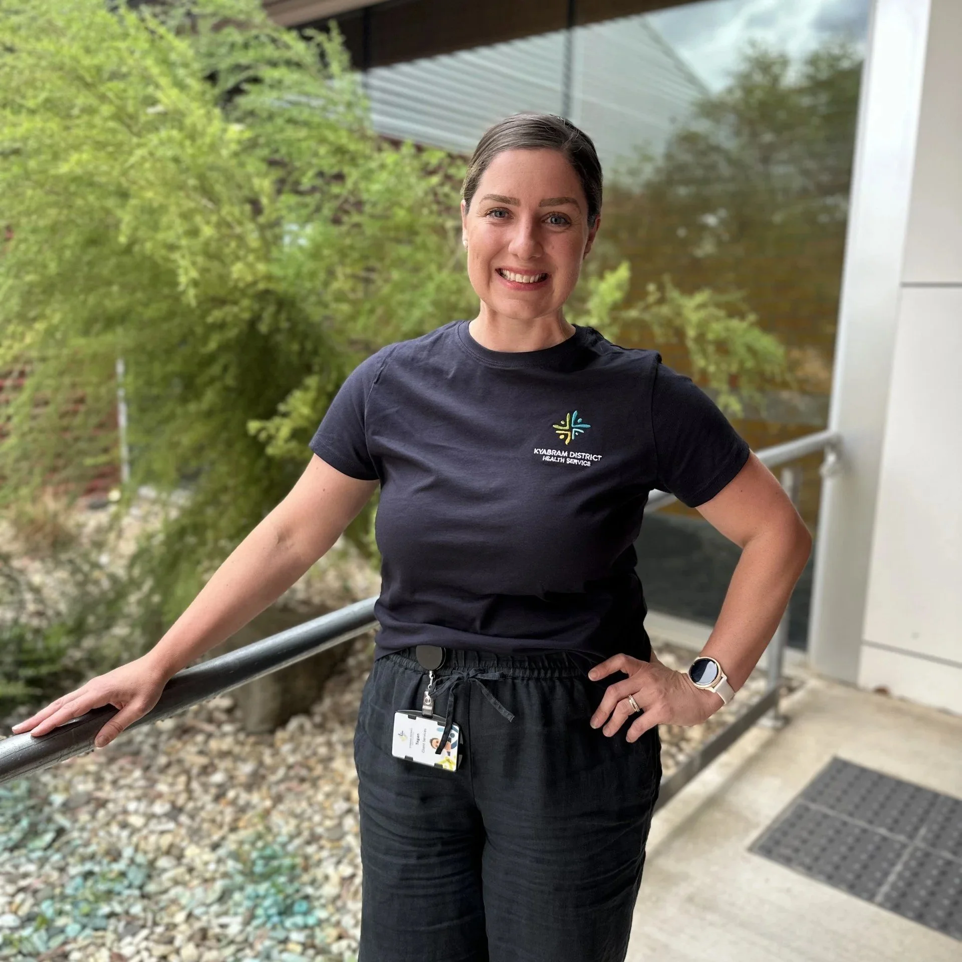 A smiling woman with brown hair tied back standing outdoors. She is wearing a black t-shirt with a colorful logo and the words 'Kyabram District Health Service' on it, along with a smartwatch on her left wrist. The background features green foliage, rocks, and a modern building with glass and metal siding.