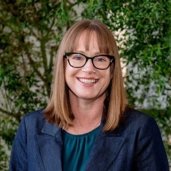 A woman with shoulder-length reddish-brown hair and glasses, smiling, wearing a navy blazer and a teal top, with a green leafy background.