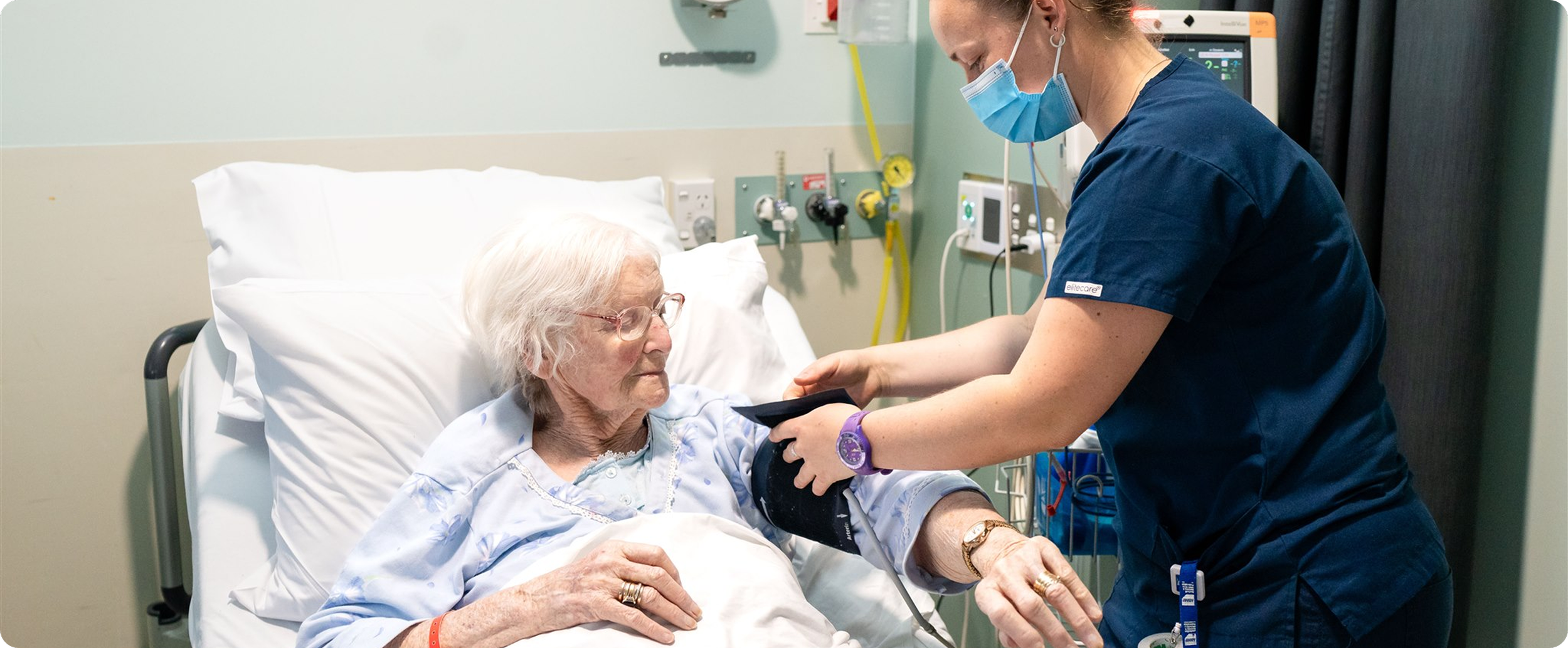 A nurse checks the blood pressure of an elderly woman lying in a hospital bed.