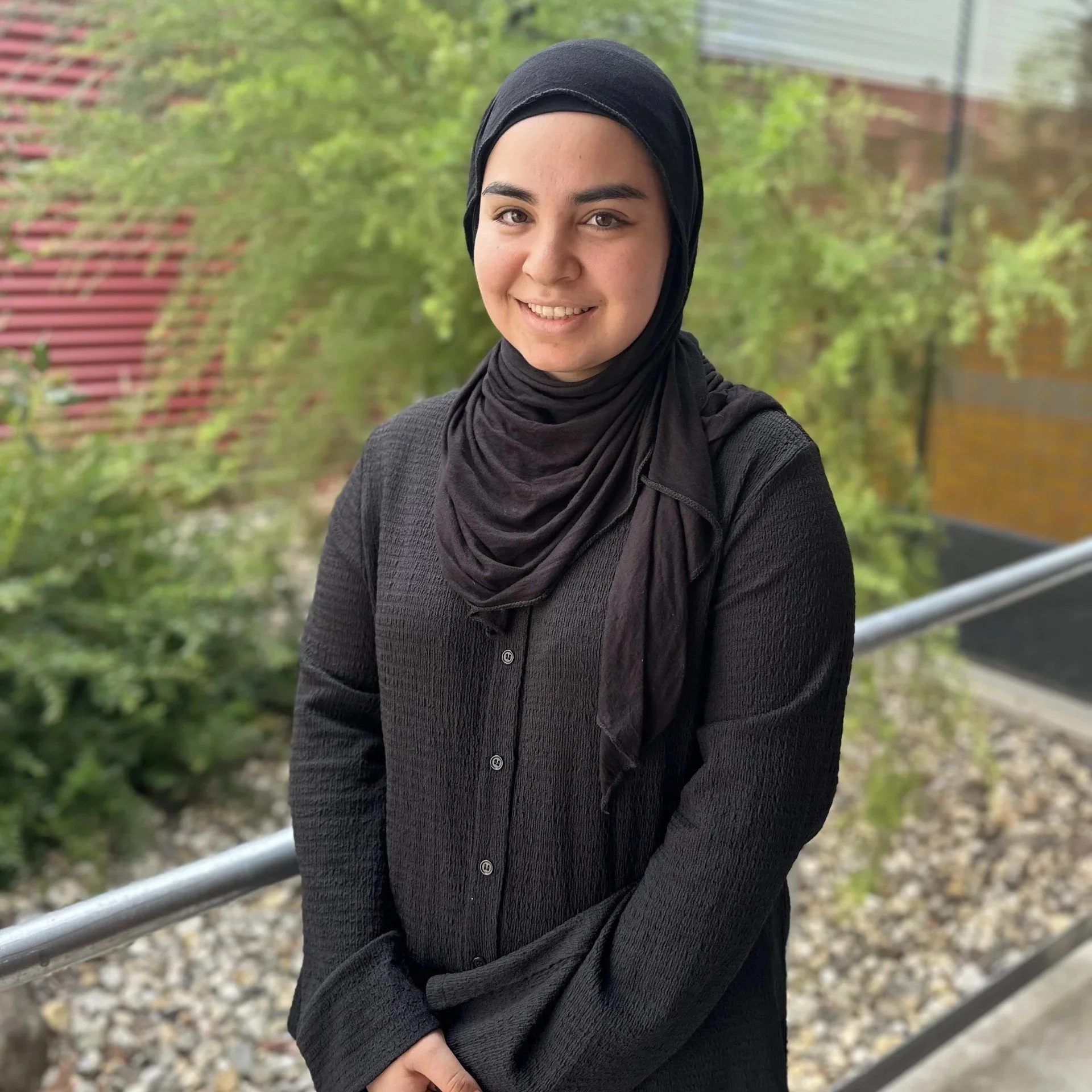 Young woman wearing a black hijab and black sweater, smiling, standing outdoors near a metal railing with green trees and building in the background.