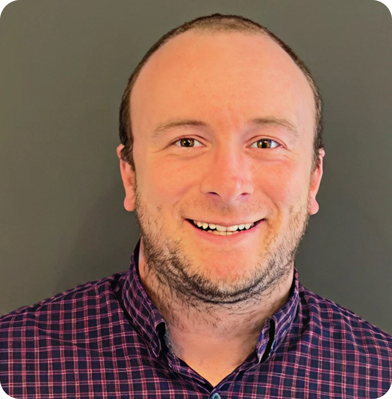 A man with short brown hair, wearing a purple and pink checkered shirt, smiling in front of a plain gray background.