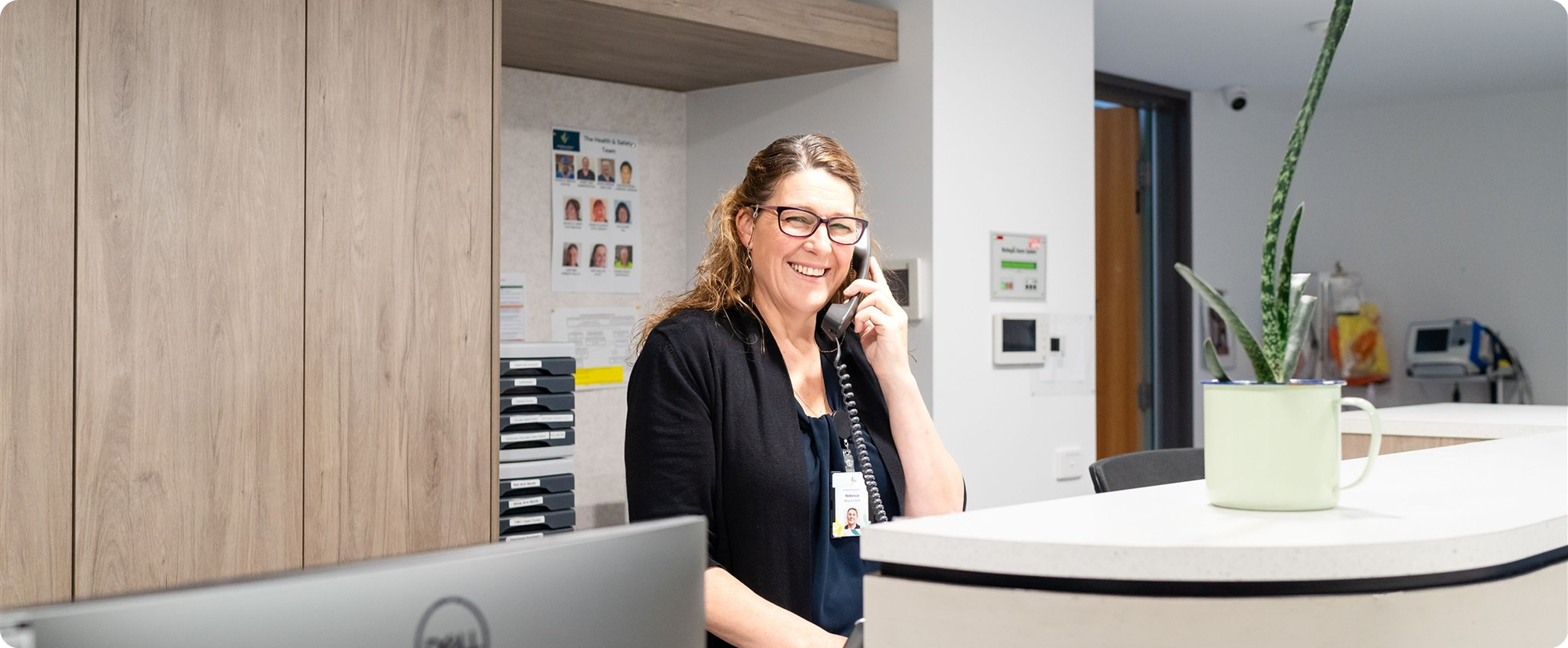 A woman with curly hair, glasses, and a black cardigan smiling while talking on a landline phone behind a white curved counter in a medical office or reception area, with a potted snake plant on the counter.