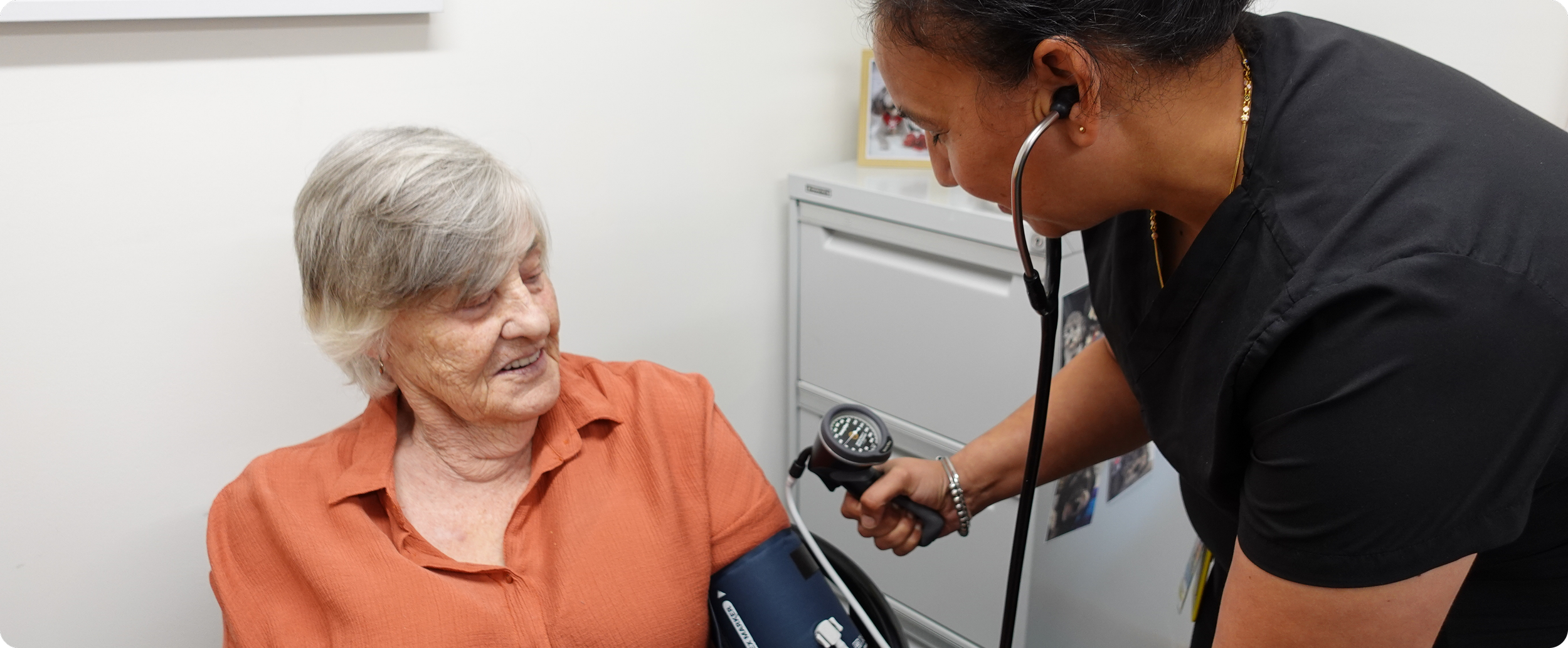 A healthcare worker using a stethoscope to check an elderly woman's blood pressure in a medical office.