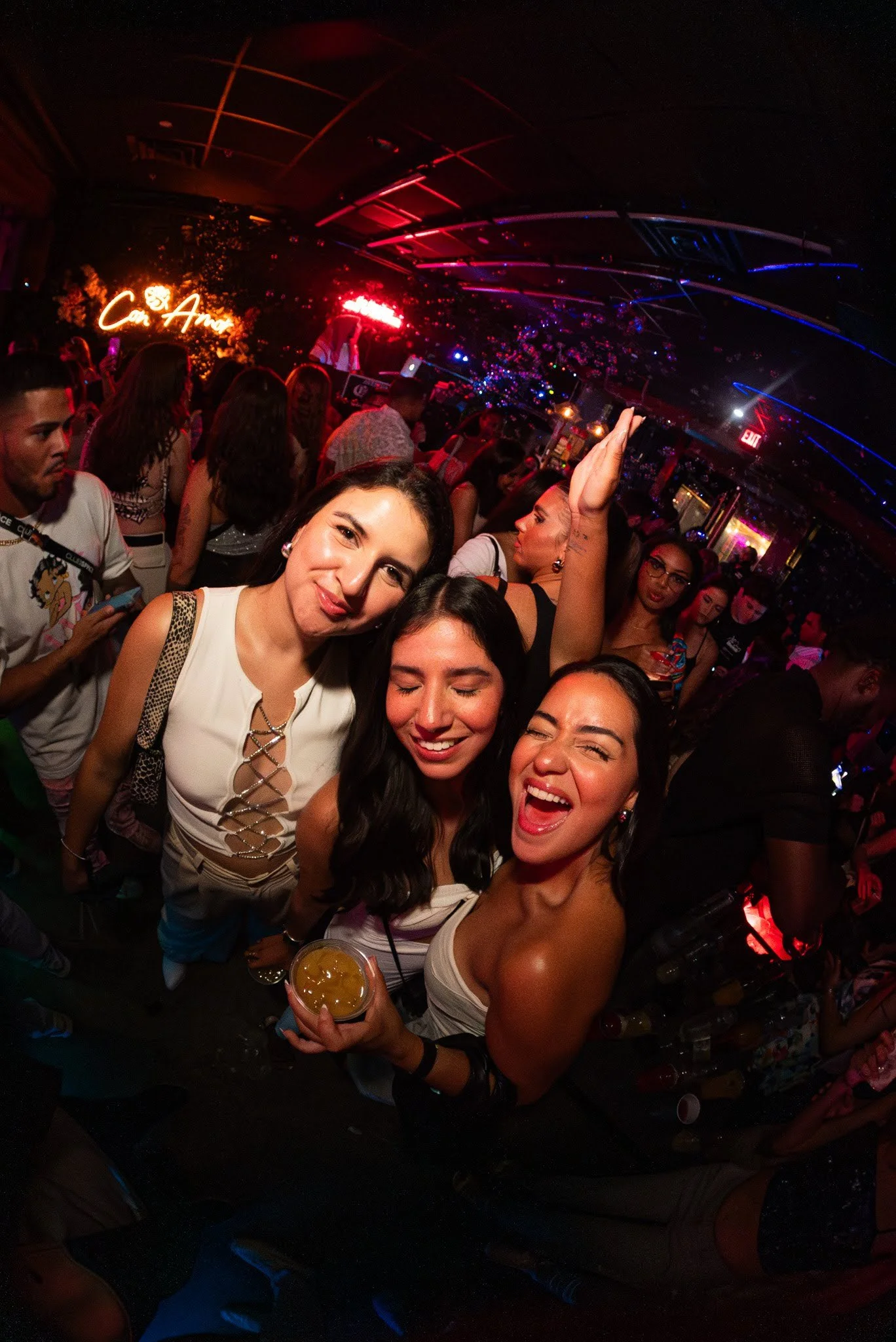 Three women celebrating at a lively nightclub, smiling and holding drinks, with other partygoers and colorful neon lighting in the background.