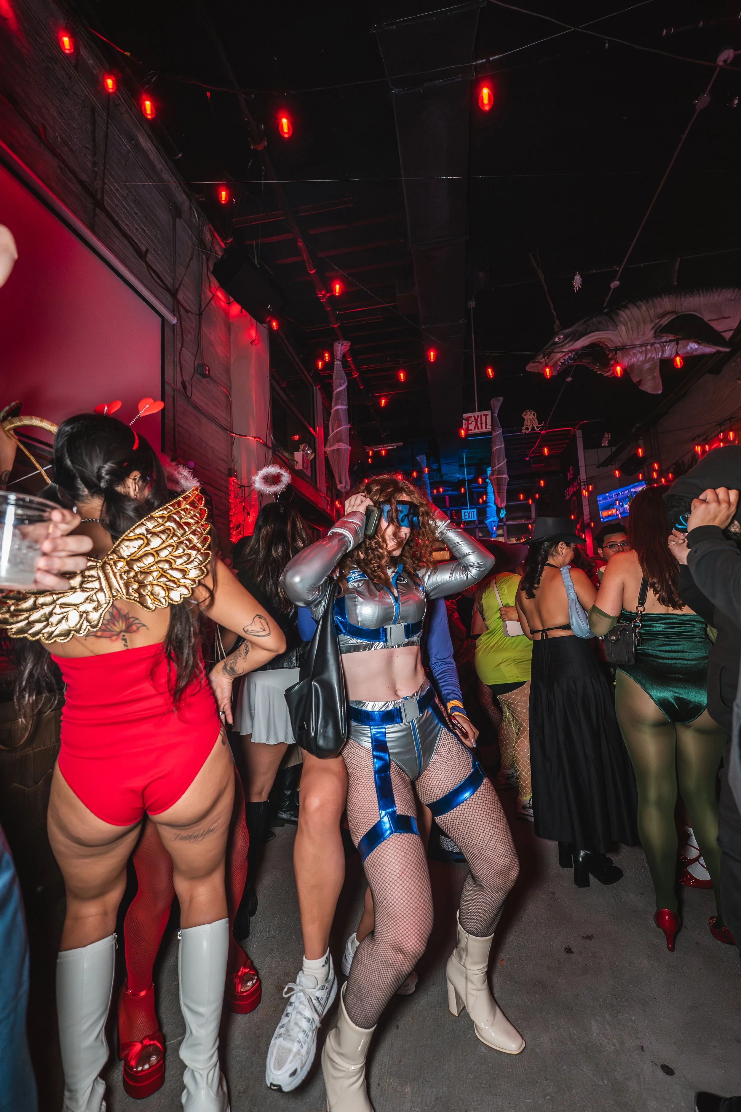 People dressed in colorful and elaborate costumes at a party or event with Halloween decorations, including hanging spider webs and a large fish decoration on the ceiling.