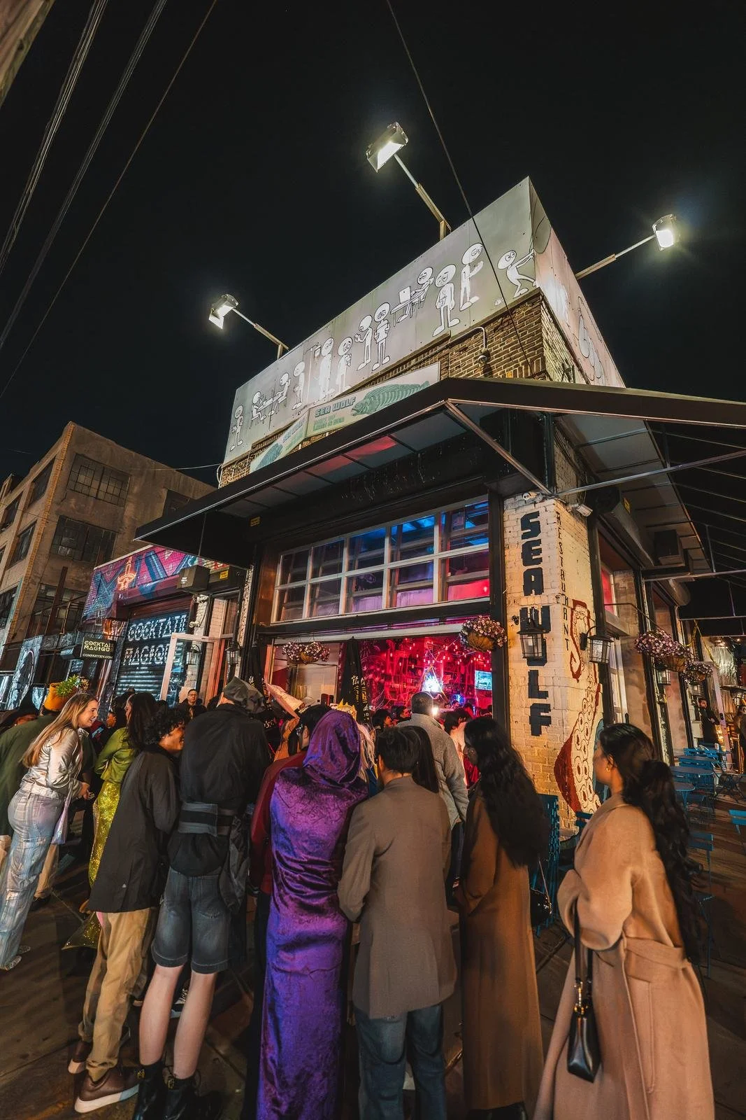 Costumed guests lined up outside the Sea Wolf entrance at night for Monstera Nightlife in Bushwick, Brooklyn, Halloween 2025