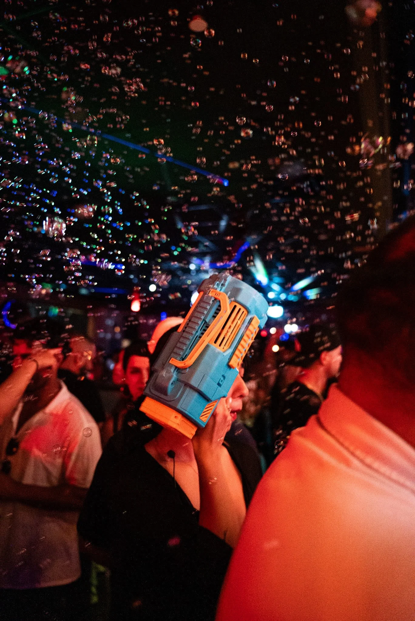 A person wearing a novelty hat resembling a gun at a lively party with colorful lights and confetti.