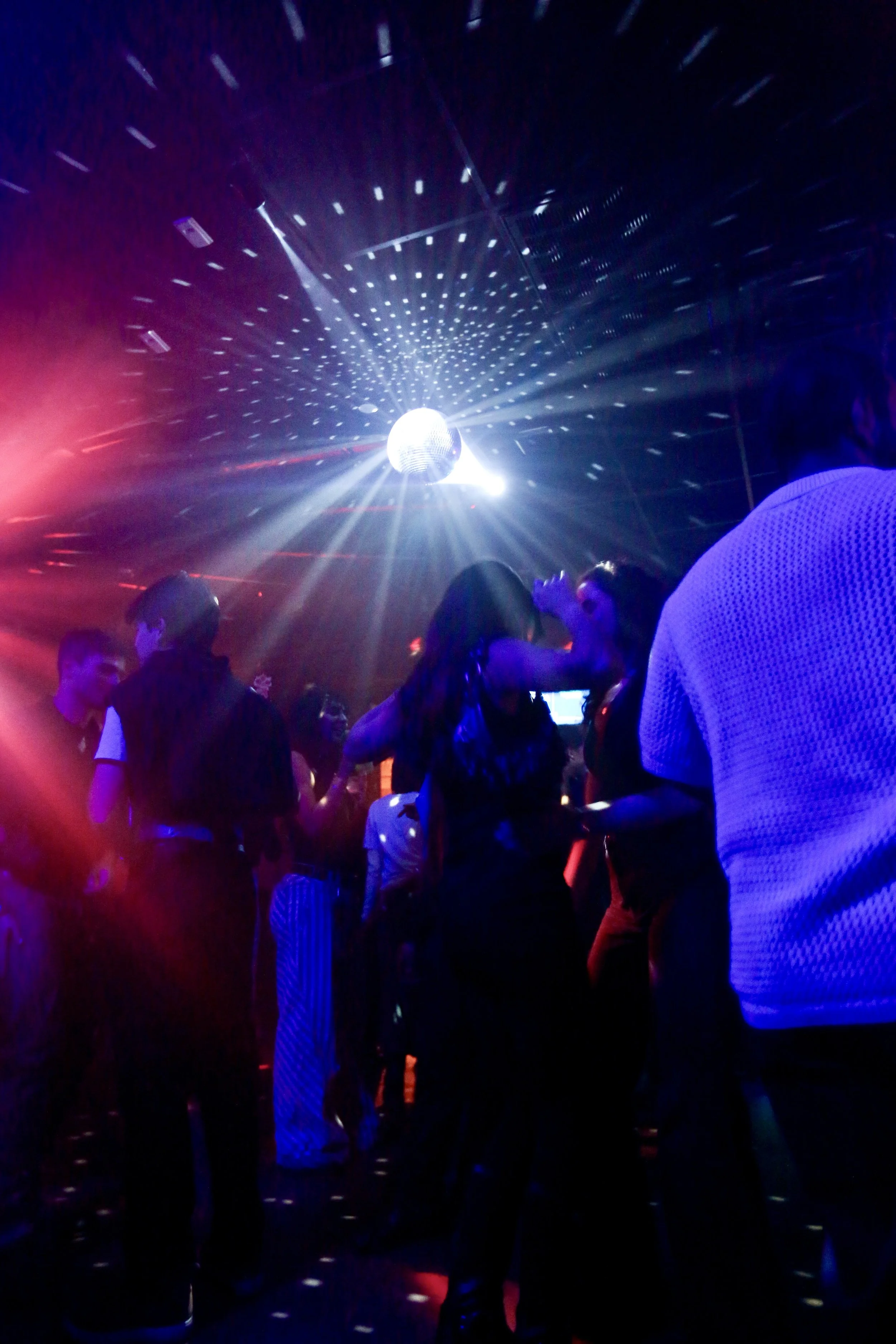 People dancing in a nightclub with colorful lights and a disco ball overhead.