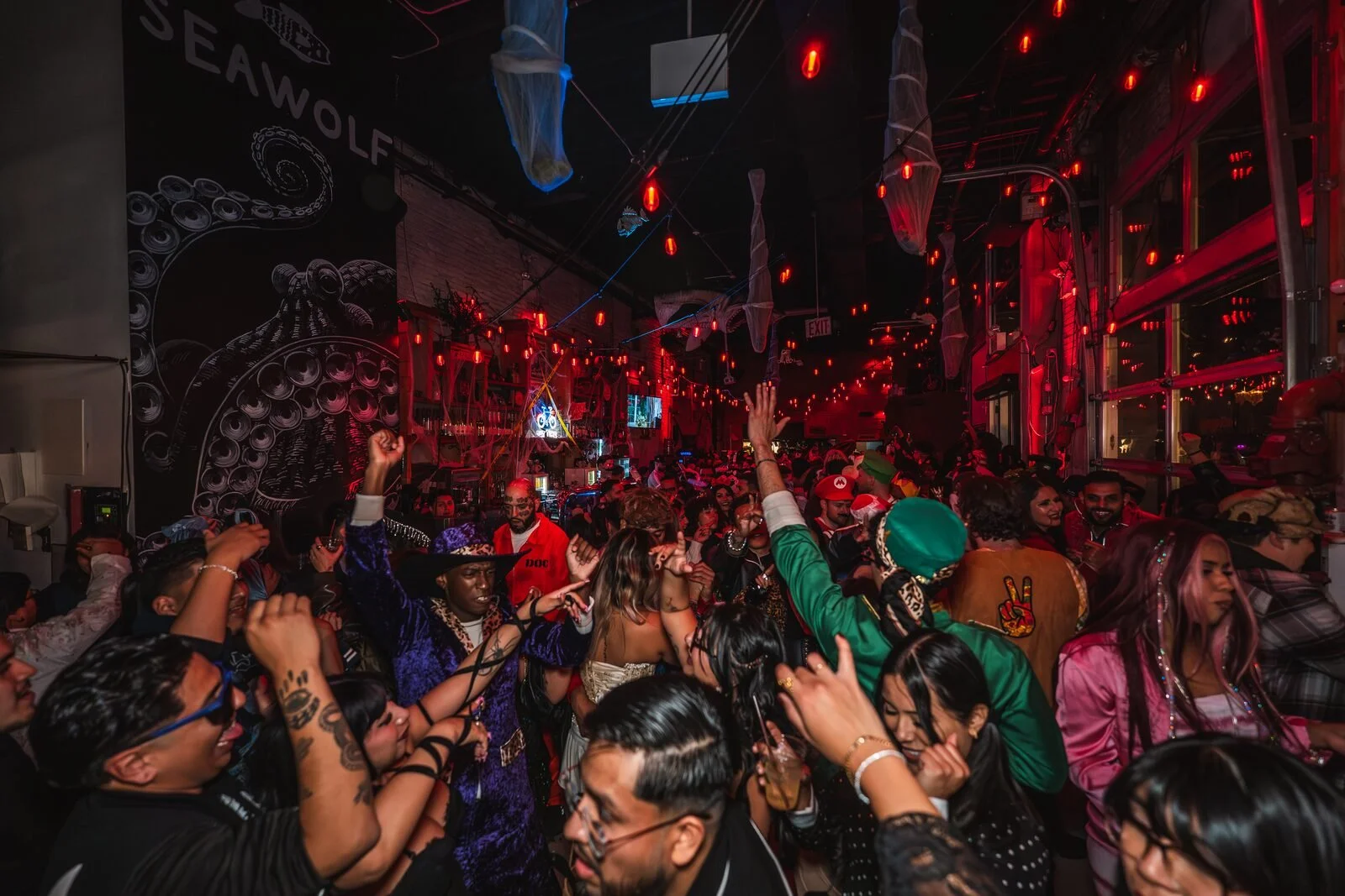 Crowd with hands raised under red lights beside the mural at a reggaeton party at Sea Wolf in Bushwick, Brooklyn