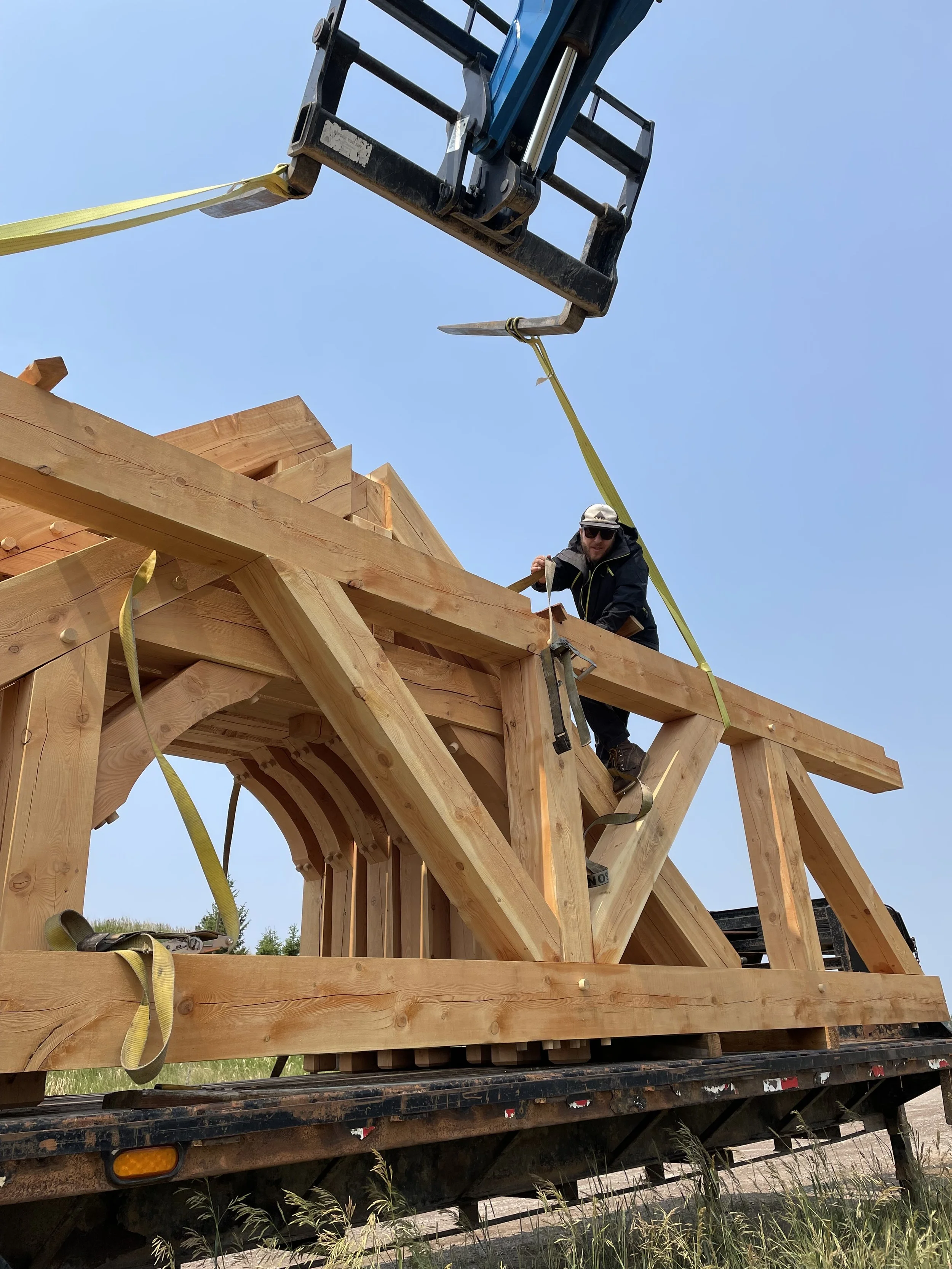 A construction worker wearing a hat, sunglasses, and black clothing is standing on a wooden structure, using a strap to secure a large wooden beam. Above, a blue crane with a metal fork is lifting a strap.