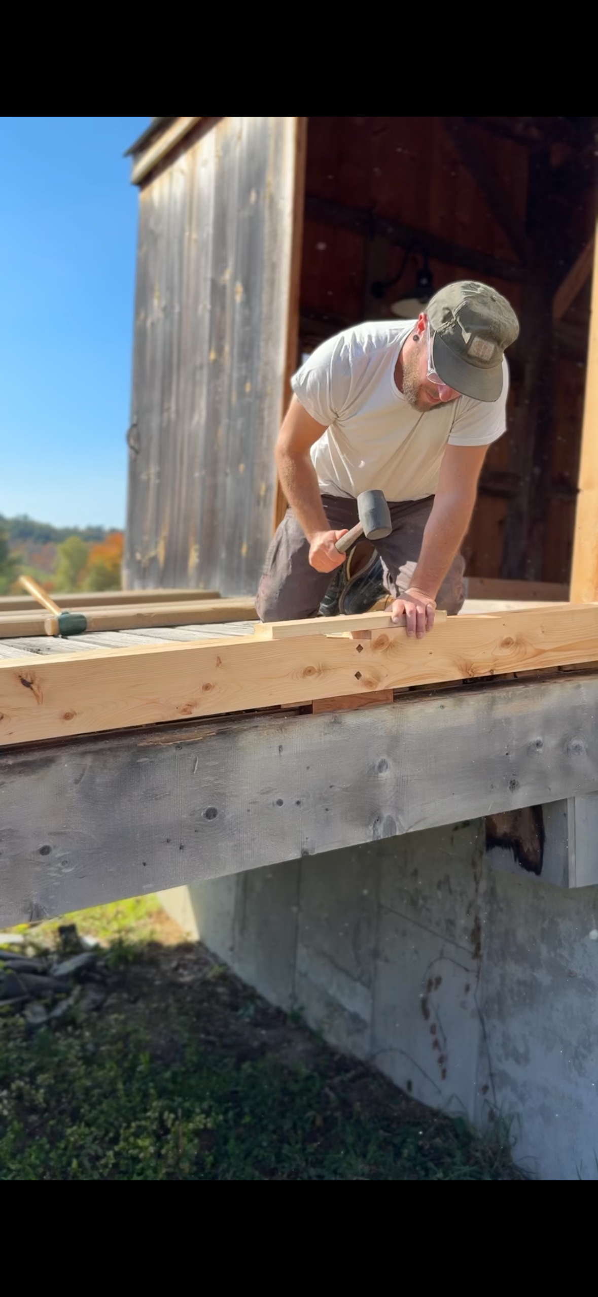 A man wearing a gray cap, white t-shirt, and glasses works on a wooden structure on a sunny day, using a hammer to secure a plank of wood.