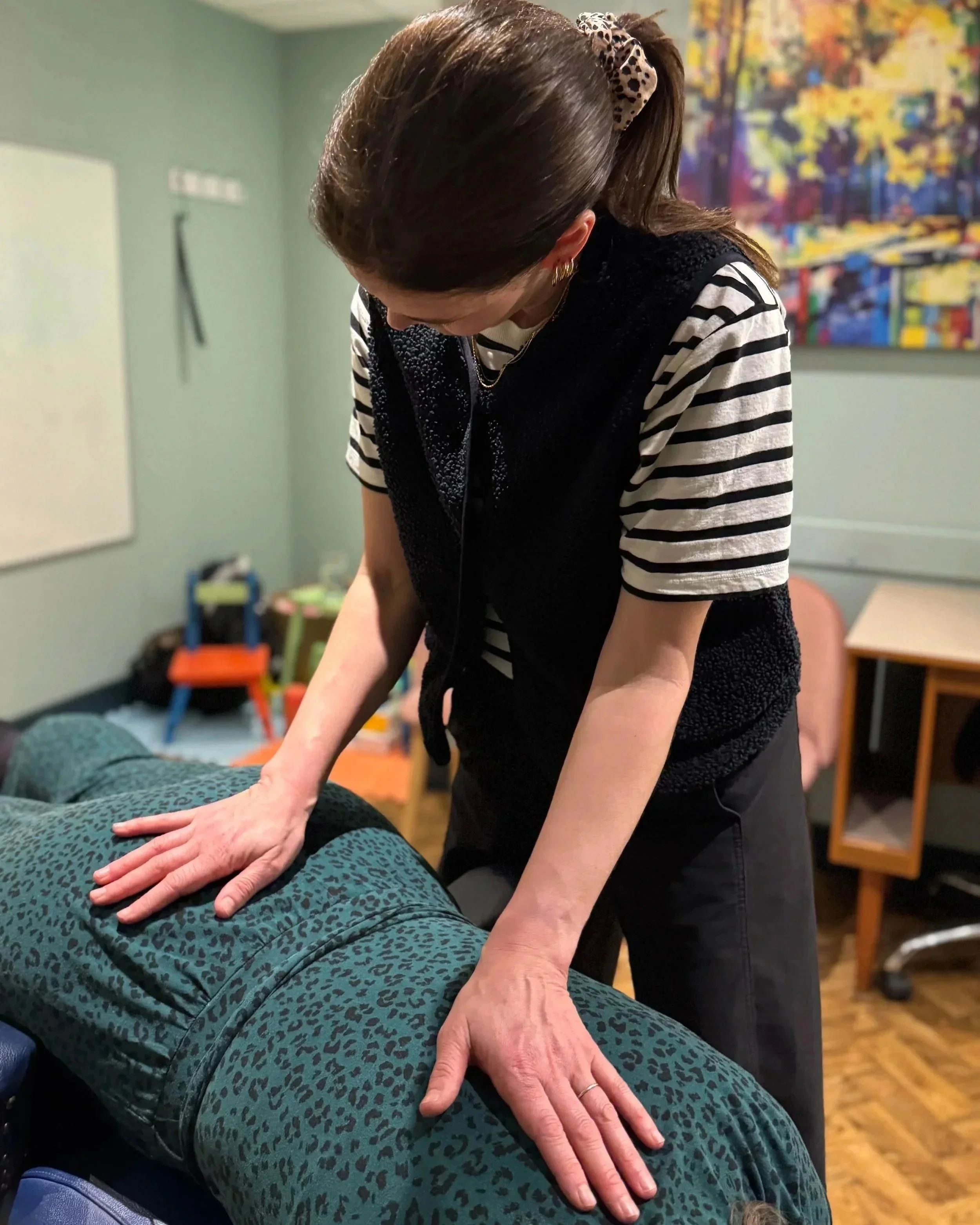 A woman providing a chiropractic adjustment to a person lying face down on a massage table in a room with colorful artwork on the wall.