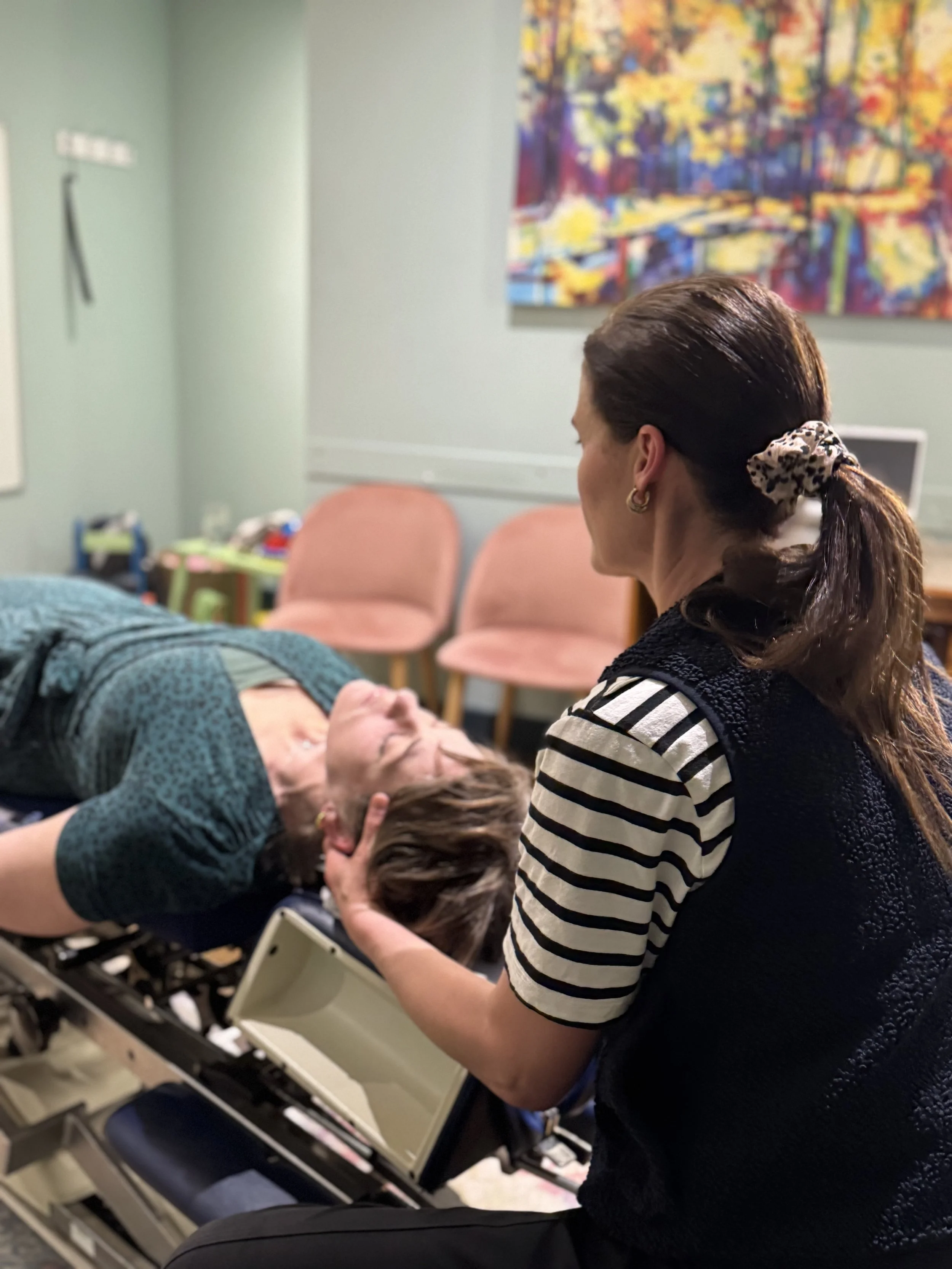 A woman lying on a chiropractic bench with her eyes closed, while a female chiropractor, or healthcare professional gently holds her head and is providing care.